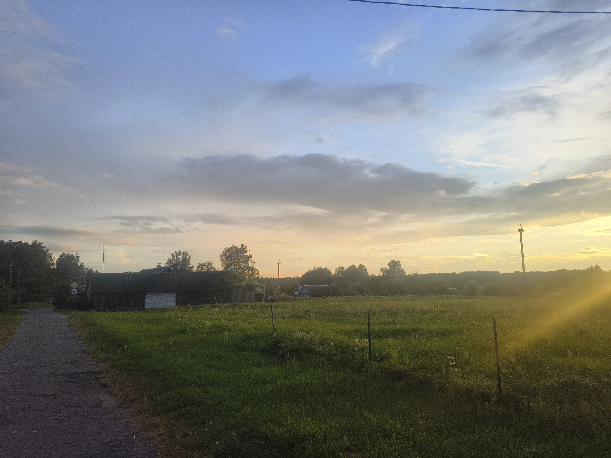 A low-angle, medium shot captures a rural scene in Kruglitski Sel'ski Sovet, Belarus, at dusk. The sky is a gradient of blue and yellow, with scattered clouds, and a power line runs horizontally across the top. A gravel road, leading from the lower left corner, cuts diagonally across the frame and disappears behind a dark, wooden structure on the left. This building, with a white-covered opening, appears to be an old barn or shed.
The foreground is dominated by a lush, green field, dotted with small white flowers and punctuated by two thin, dark fence posts. In the mid-ground, beyond the field, a dense line of trees forms a natural boundary, with several small buildings and structures visible within the treeline. To the right, a tall, dark utility pole stands in the field, with sunlight flaring from the right side of the image, creating a bright, luminous effect. The overall atmosphere is peaceful and serene, indicative of a quiet evening in the Belarusian countryside.