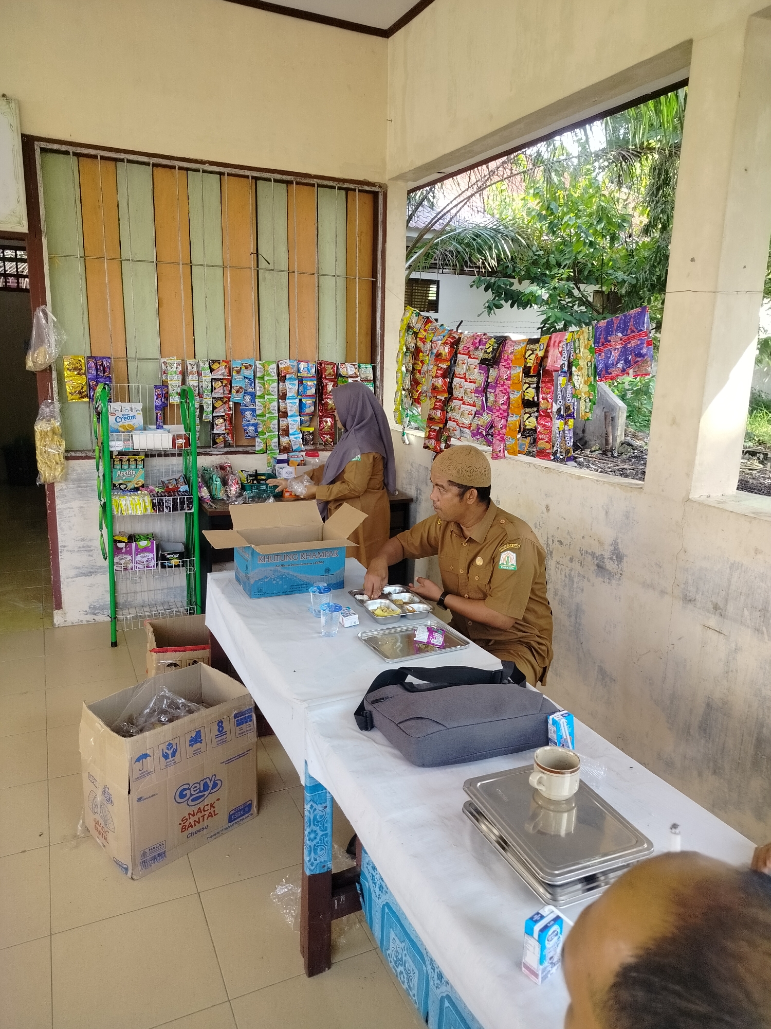 The image depicts a simple outdoor food stall or canteen, likely in Purwodadi, Indonesia. Two individuals, dressed in official uniforms, are present. One is a woman wearing a hijab and a brown uniform, attending to items behind a counter where various packaged snacks are displayed on shelves and hung from a line. The other is a man, also in a brown uniform and a cap, seated at a table covered with a white tablecloth. He is in the process of eating from a metal tray containing several small dishes of food. On the table, there are also cups, small cartons of milk, and a grey bag. In the foreground, to the left, a large cardboard box labeled "Gery SNACK BANTAL Cheese" is visible on the tiled floor. The stall appears to be in an open-air setting, with trees and greenery visible in the background through an opening. The overall impression is that of a casual, utilitarian food service area, possibly associated with a government office or public facility given the uniforms.