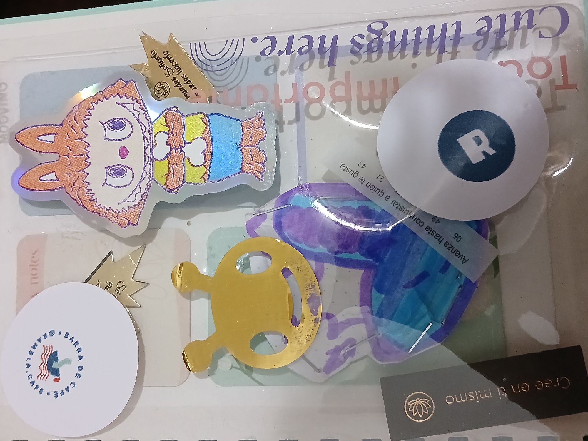 The image presents a flat lay of various decorative and functional items, including stickers, cards, and a tag, arranged under a clear plastic cover, likely part of a planner or journal. The collection suggests a personal assortment of small mementos or motivational items.
In the upper left, a holographic sticker depicts a cartoon character with bunny ears, an orange-brown face, purple eyes, a small pink nose, and a yellow top with bone patterns, paired with blue pants. Above this character, a small golden, spiky-edged piece of paper displays the Spanish text "Puedes hacerlo, sueña" (You can do it, dream!).
To the right, a white circular sticker features a dark blue circle containing a white letter "R". Below it, a transparent piece of material with abstract purple and blue watercolor-like patterns has a small label reading "Avanza hasta conquistar a quien te gusta." (Move forward until you win over the one you like.), followed by numbers "21 43" and "06 49".
In the bottom left, a white circular sticker showcases a stylized red and blue wavy logo, encircled by the text "CAFE • BARRA DE • @RAMBLA.C". Centrally located is a metallic gold-colored cutout, possibly resembling a minimalist alien head or a stylized steering wheel.
At the bottom right, a black rectangular tag or sticker is visible, featuring a golden lotus flower symbol and the Spanish phrase "Cree en ti mismo" (Believe in yourself). The background shows faint text from what appears to be planner pages, with visible phrases like "Cute things here.", "Important", and "notes". The prevalence of Spanish text throughout these items aligns with the given location context of Buenos Aires, Argentina.
