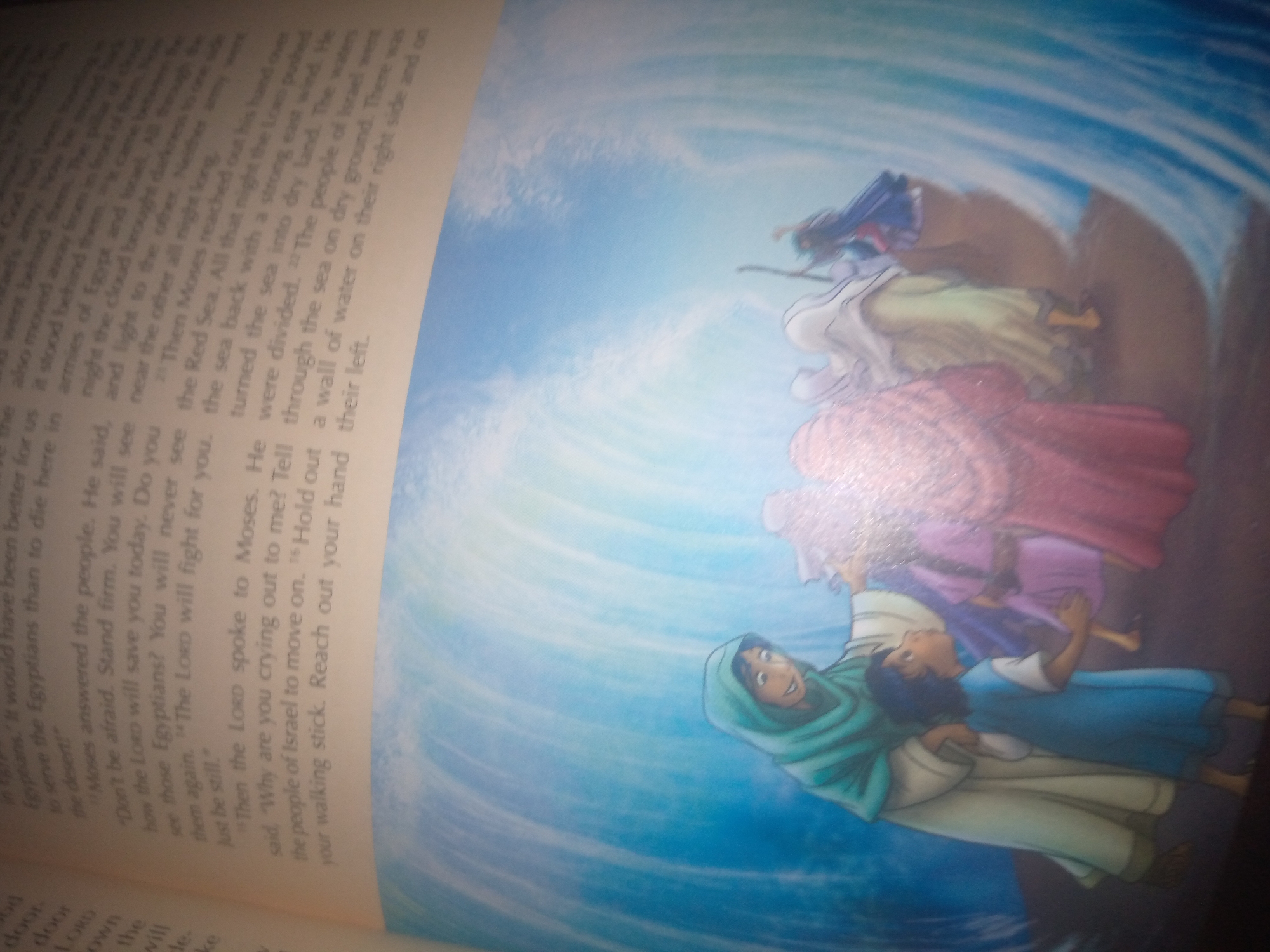 The image is a close-up, angled photograph of an open book. The book's pages display text on the left and an illustration on the right, set against a backdrop of swirling blue and white patterns that suggest water. The text on the left page is partially visible and appears to be from a religious scripture, with numbered verses. The illustration on the right depicts several figures in stylized, cartoonish attire. One figure, wearing a green headdress, is prominent in the foreground, looking upwards with a smile. Behind this figure, other figures are shown, with a central figure appearing to be parting the blue water with a stick. The scene is brightly lit, with some glare visible on the glossy illustration. The image does not contain any elements that suggest a specific location in Jalingo, Nigeria, or any urban setting.