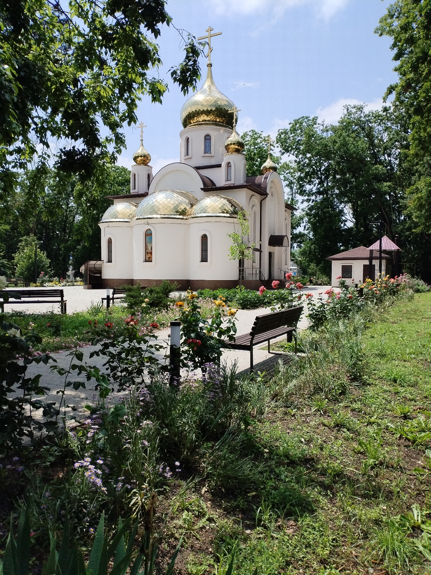 The image captures a white church with golden domes, set against a backdrop of lush green trees and a blue sky with scattered clouds. In the foreground, a paved walkway curves through a garden filled with various flowers, including roses and lavender. Two dark wooden benches are placed along the walkway, offering a place to rest and admire the scenery. To the right, a small, brown, shed-like structure with a red-tiled roof stands near the flowerbeds. The scene evokes a sense of tranquility and natural beauty, with the church serving as a focal point in the serene environment of Vinogradny, Russia.