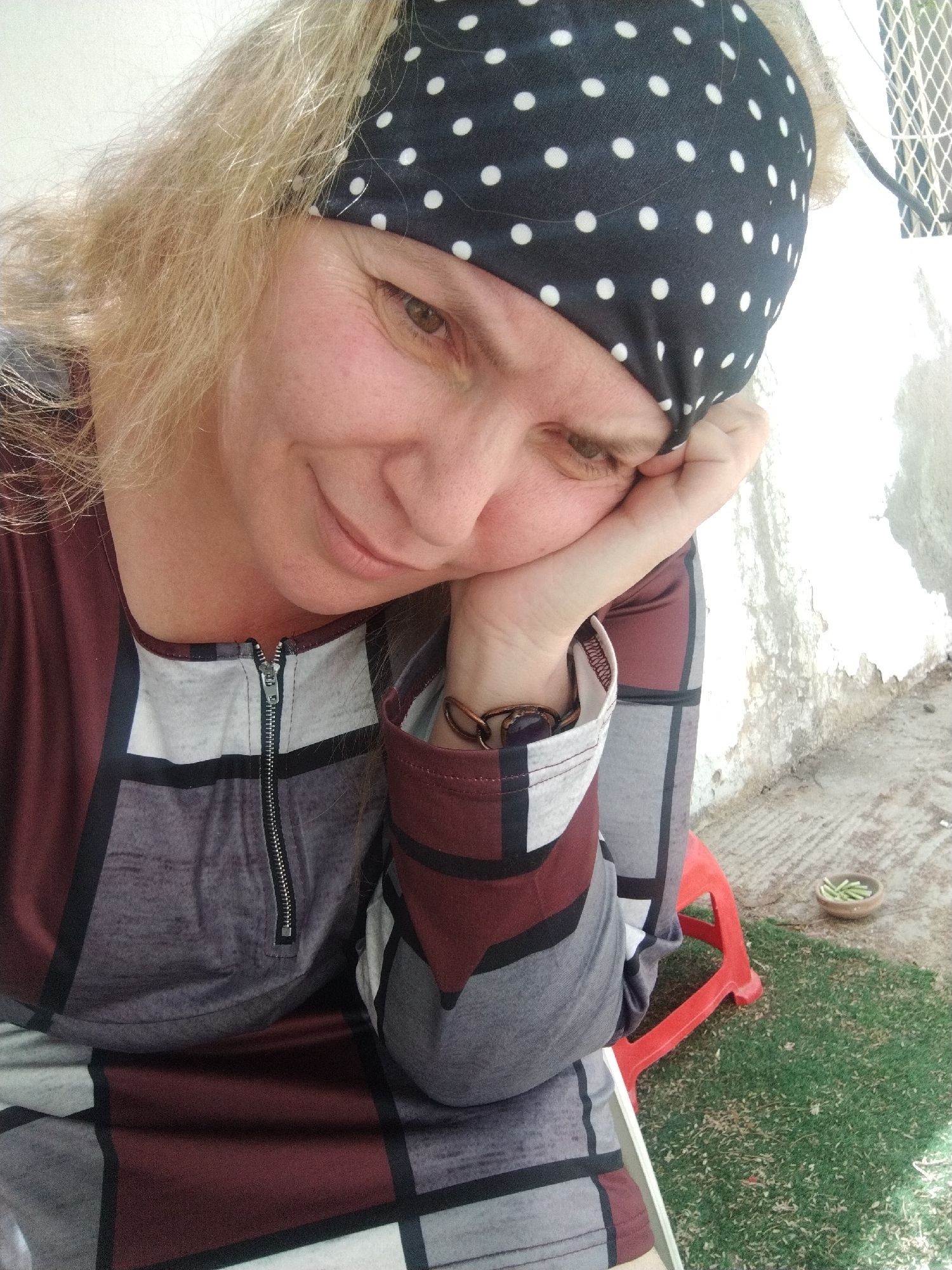 A woman is sitting with her head resting on her left hand. She is wearing a black and white polka dot headband and a long-sleeved dress with a geometric pattern of maroon, gray, black, and white. A silver zipper is partially visible at the neckline. She is wearing a bracelet on her left wrist. The woman appears to be on a patio or outdoor area, with a small red plastic stool visible behind her. A small dish with green pieces is on the ground. The location is Al Mas'oudi, United Arab Emirates.