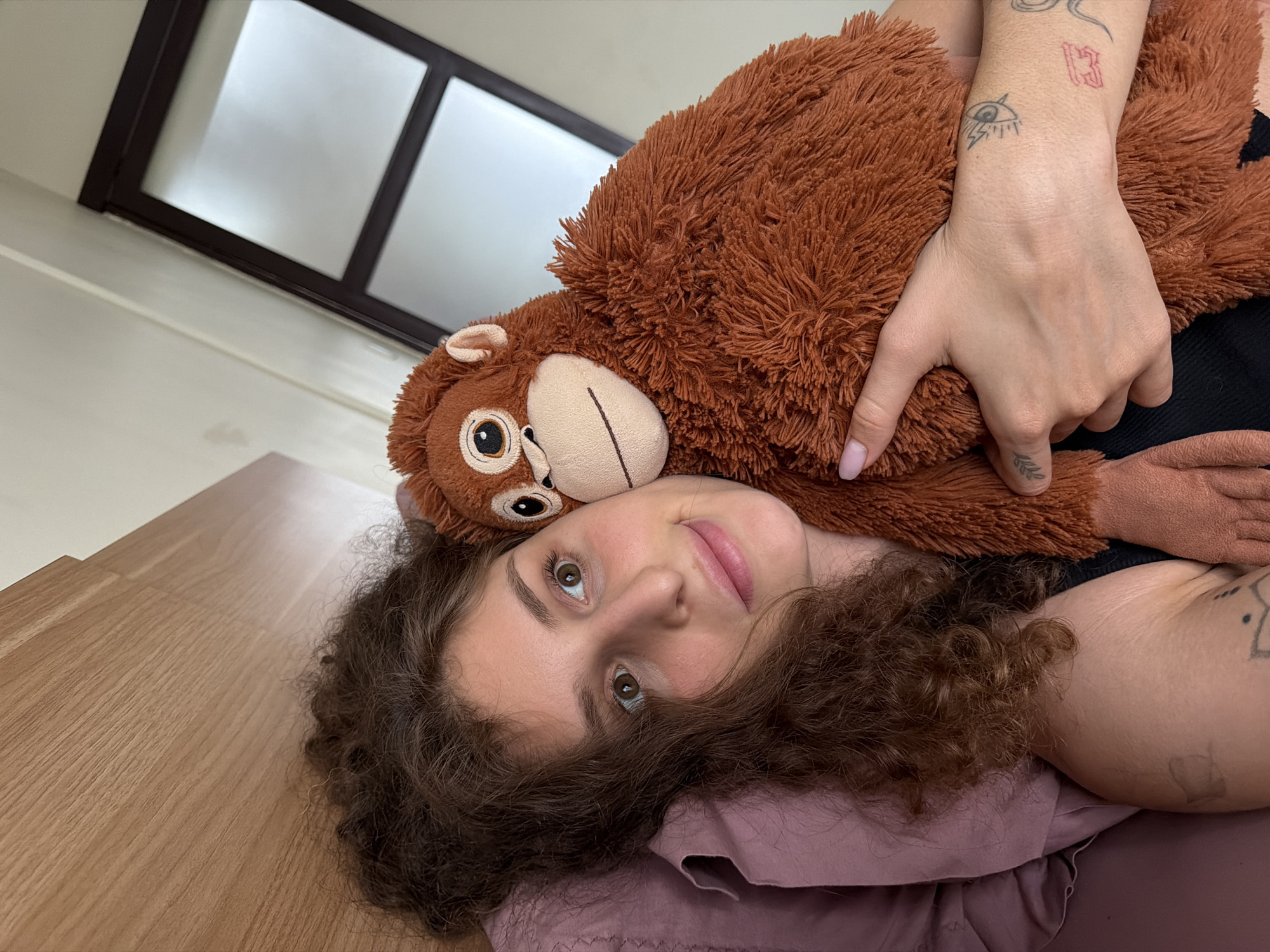 A young woman with curly brown hair lies in bed with a plush monkey toy. The woman is looking at the camera and holding the monkey. The room appears to be a bedroom, with a wooden headboard visible behind the woman. A partially obscured window with frosted glass and dark framing is visible in the background. The image was taken in North Kuta, Indonesia.