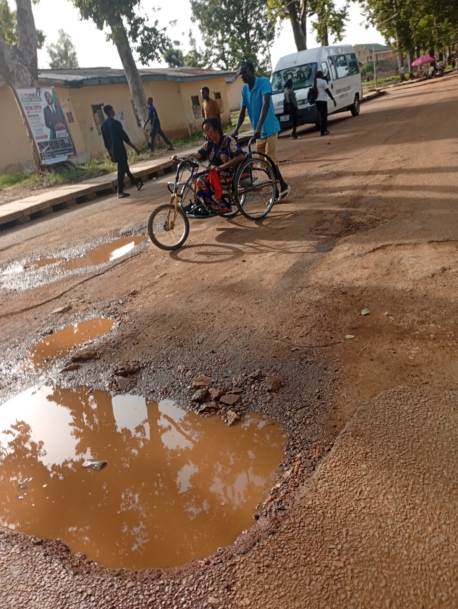 In Rafin Par, Nigeria, a man in a blue shirt and khaki shorts pushes a person in a wheelchair along a poorly maintained dirt road. The road is littered with potholes filled with muddy water. The wheelchair user is wearing a dark top with a colorful pattern and a red garment draped over their legs. Behind them, other individuals are visible, including a man in a dark shirt walking towards a building on the left, and several people near a white van parked on the right side of the road. A poster advertising a music coach is affixed to the building. Trees line the road, and the sky is visible overhead. The overall scene depicts a challenging road condition and the movement of people on a street.