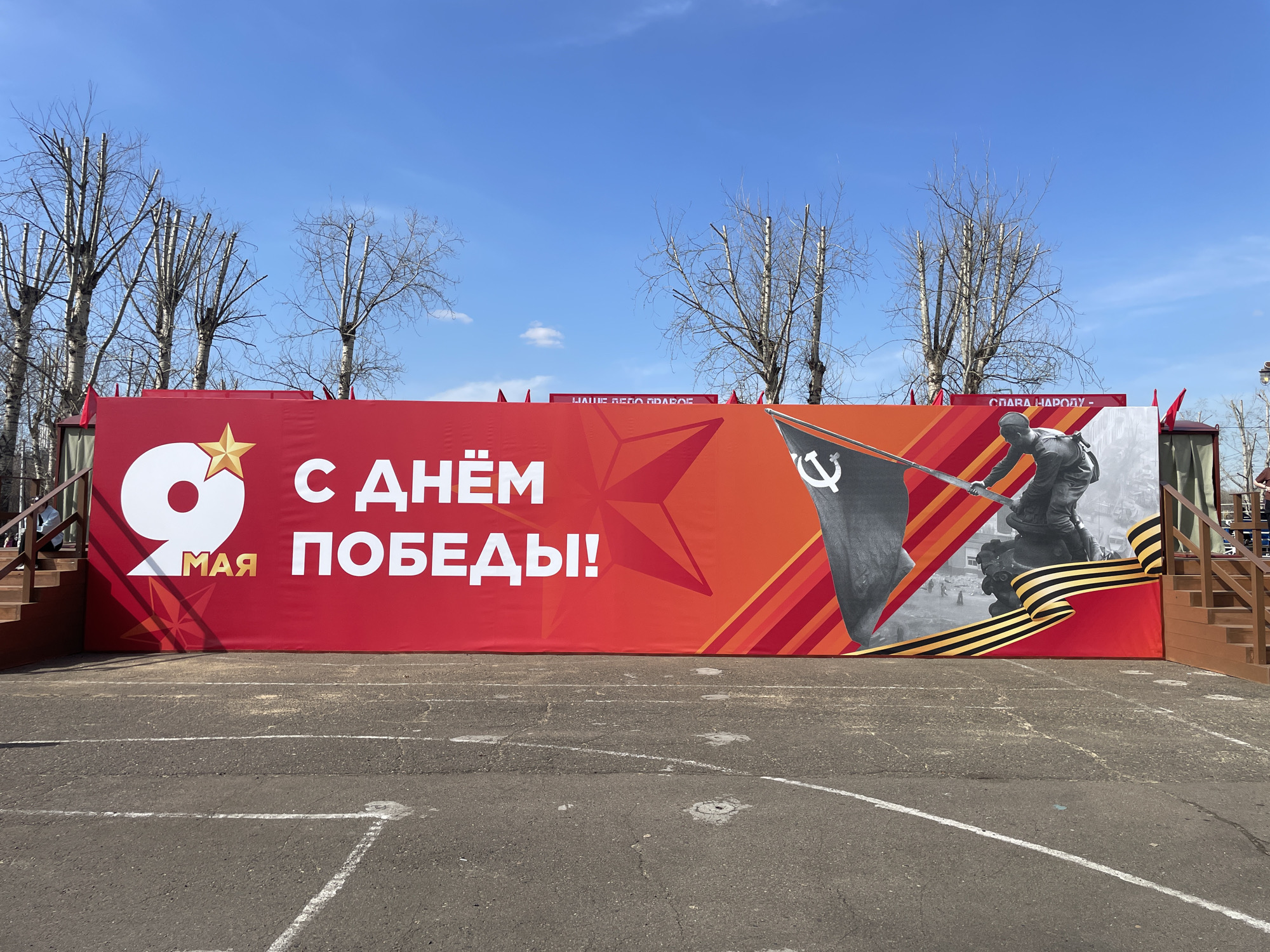 A wide, ground-level shot depicts a large red banner in front of bare trees and a clear blue sky, likely in Krasnoyarsk, Russia. The banner features the text "9 МАЯ С ДНЁМ ПОБЕДЫ!" ("May 9th, Victory Day!") in white and yellow. The banner also features an image of a Soviet soldier raising a flag with a hammer and sickle emblem. A black and orange ribbon, reminiscent of the Ribbon of Saint George, adorns the banner. The banner is set on a stage with wooden steps on either side. In front of the banner is a gray asphalt ground marked with white lines. Additional banners are visible in the background, partially obscured by the trees. People are seen standing on the wooden platforms at the sides of the main banner.