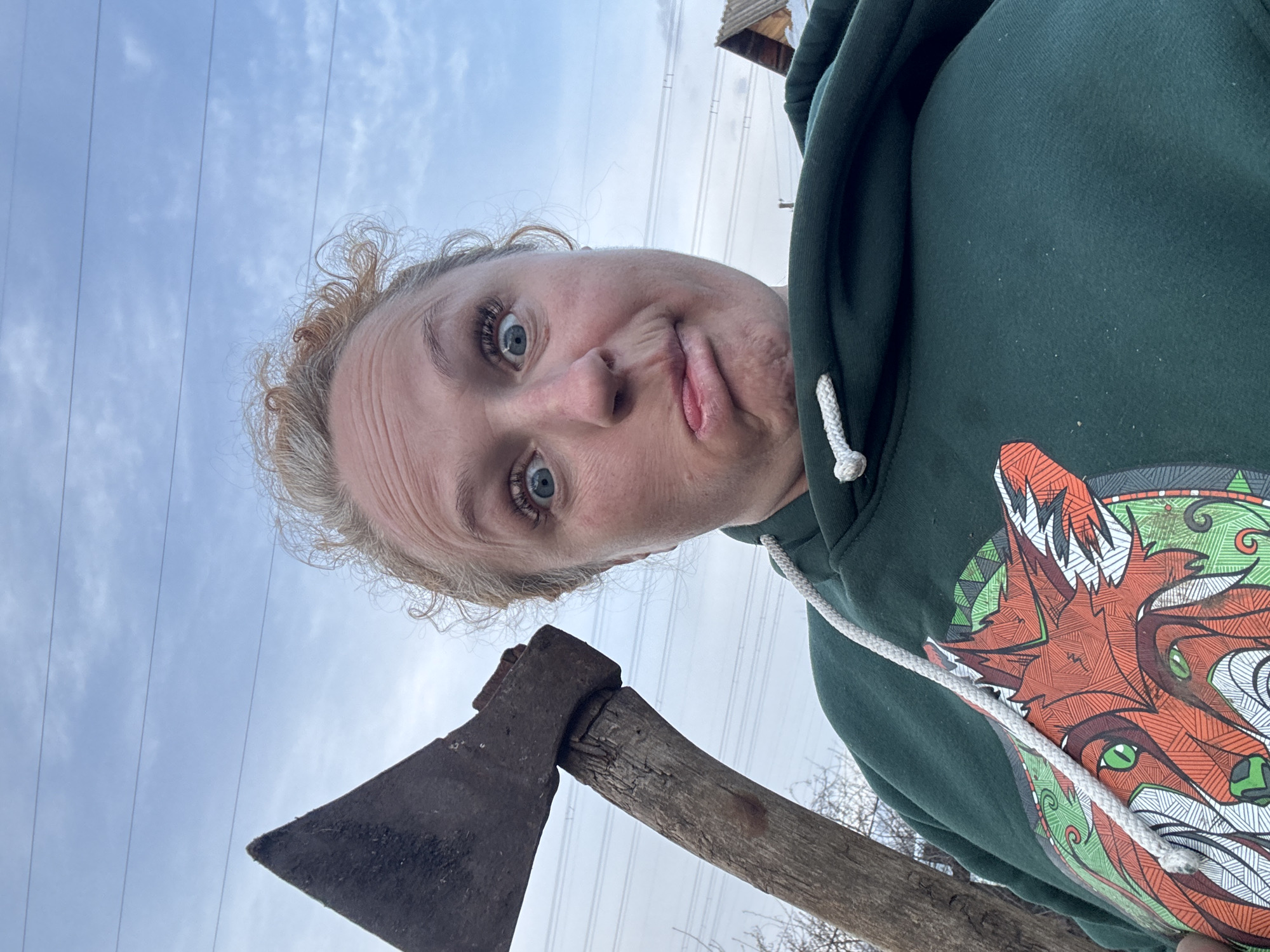 A woman is holding an axe and making a face at the camera. The sky is overcast with power lines visible. She is wearing a green hooded sweatshirt with a fox design. The scene may be located in Chaikovsky, Russia, based on information not contained in the visual content.