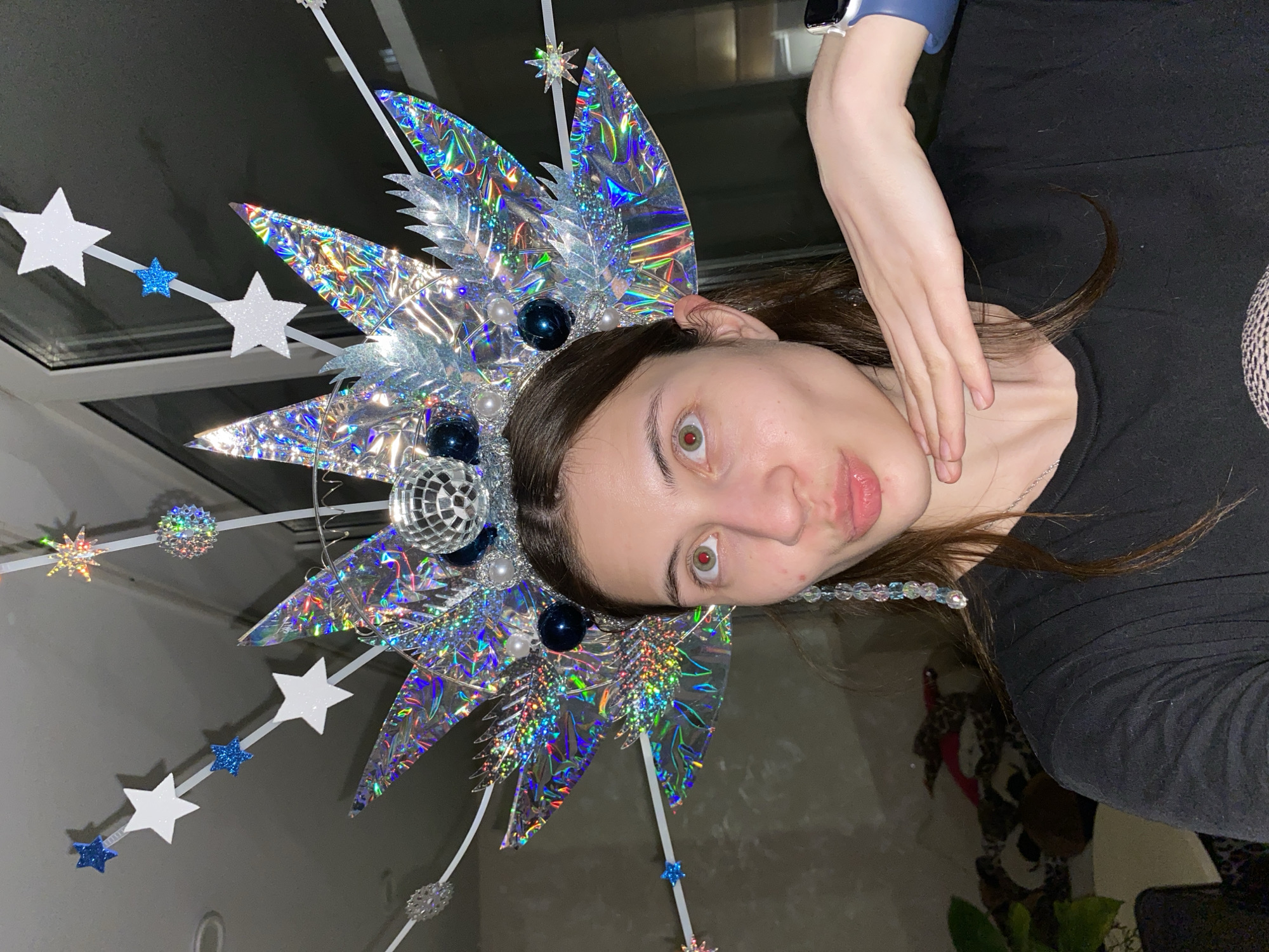A young woman with brown hair is posing for a selfie indoors. She is wearing a large, elaborate headdress adorned with holographic starbursts, disco balls, and star-shaped decorations on white rods. She is wearing a black top and has a silver necklace. Her hand is near her chin. The location appears to be inside a home, with a window visible in the background. The exact location in Votkinsk, Russia, cannot be determined from the image.