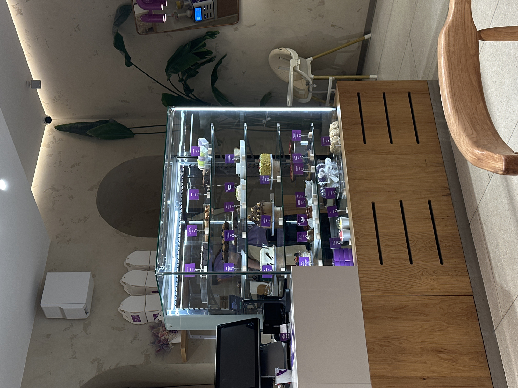 The image depicts the interior of a bakery or patisserie, likely located in Rostov-on-Don, Russia. A prominent feature is a refrigerated display case filled with various pastries and cakes. Each item has a small tag with purple accents. The display case sits on a counter next to wooden cabinetry with ventilation slits. In front of the display case is a point-of-sale system with a monitor. Adjacent to the counter is a wooden chair with a curved back.
In the background, there is a white baby high chair and a variety of plants and decorative wall fixtures. Three white take-away pastry boxes are stacked near a white dispenser. The walls are textured and off-white.