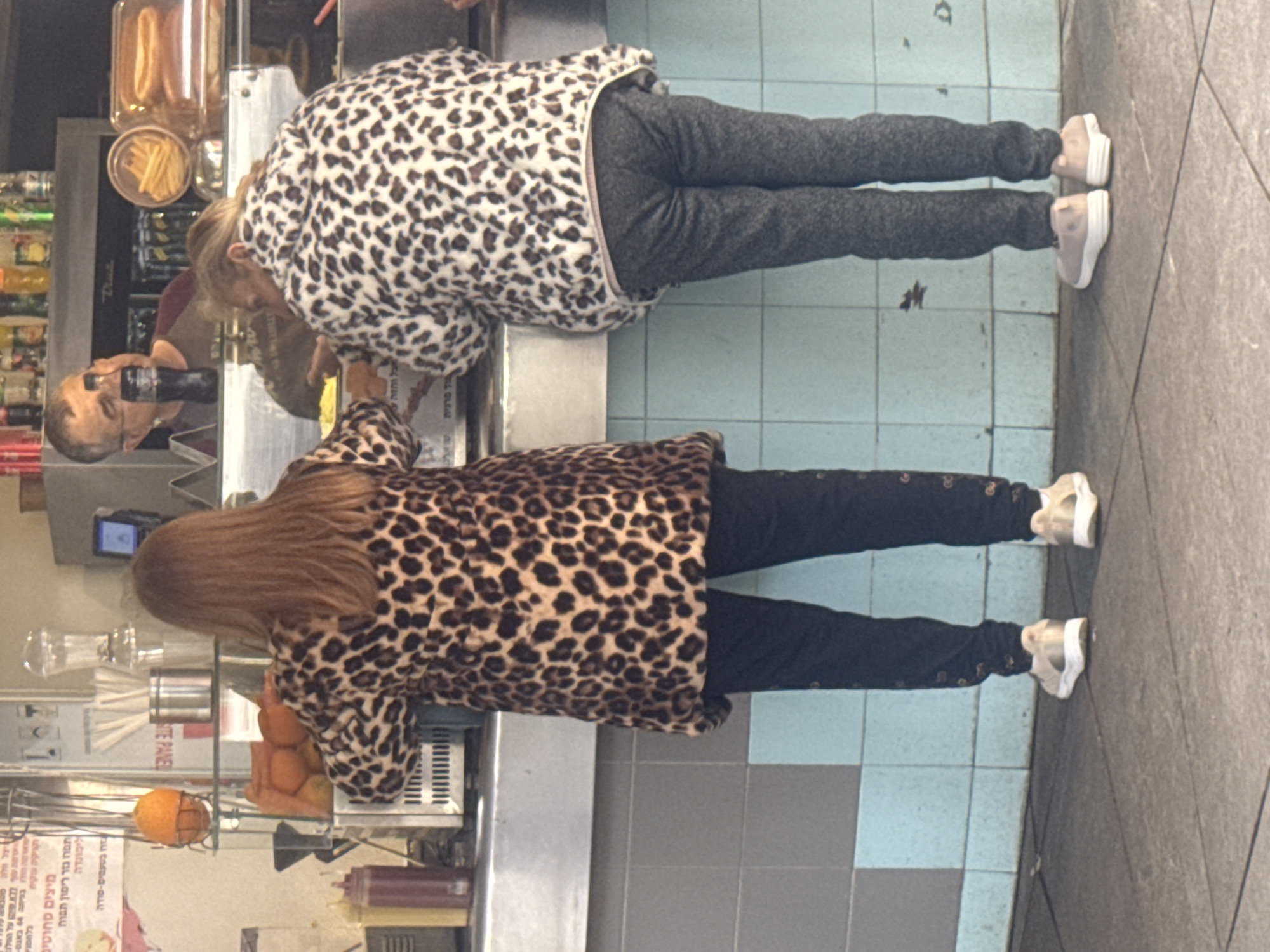 Two women wearing leopard-print jackets stand next to each other at a counter. One jacket is primarily tan with brown spots, and the other is white with brown spots. Both women wear dark pants and light-colored sneakers. A man stands behind the counter. The counter appears to be in a food establishment with beverage containers, produce, and food items visible. The floor has square tiles of varying colors. The location is Ramat Gan, Israel.