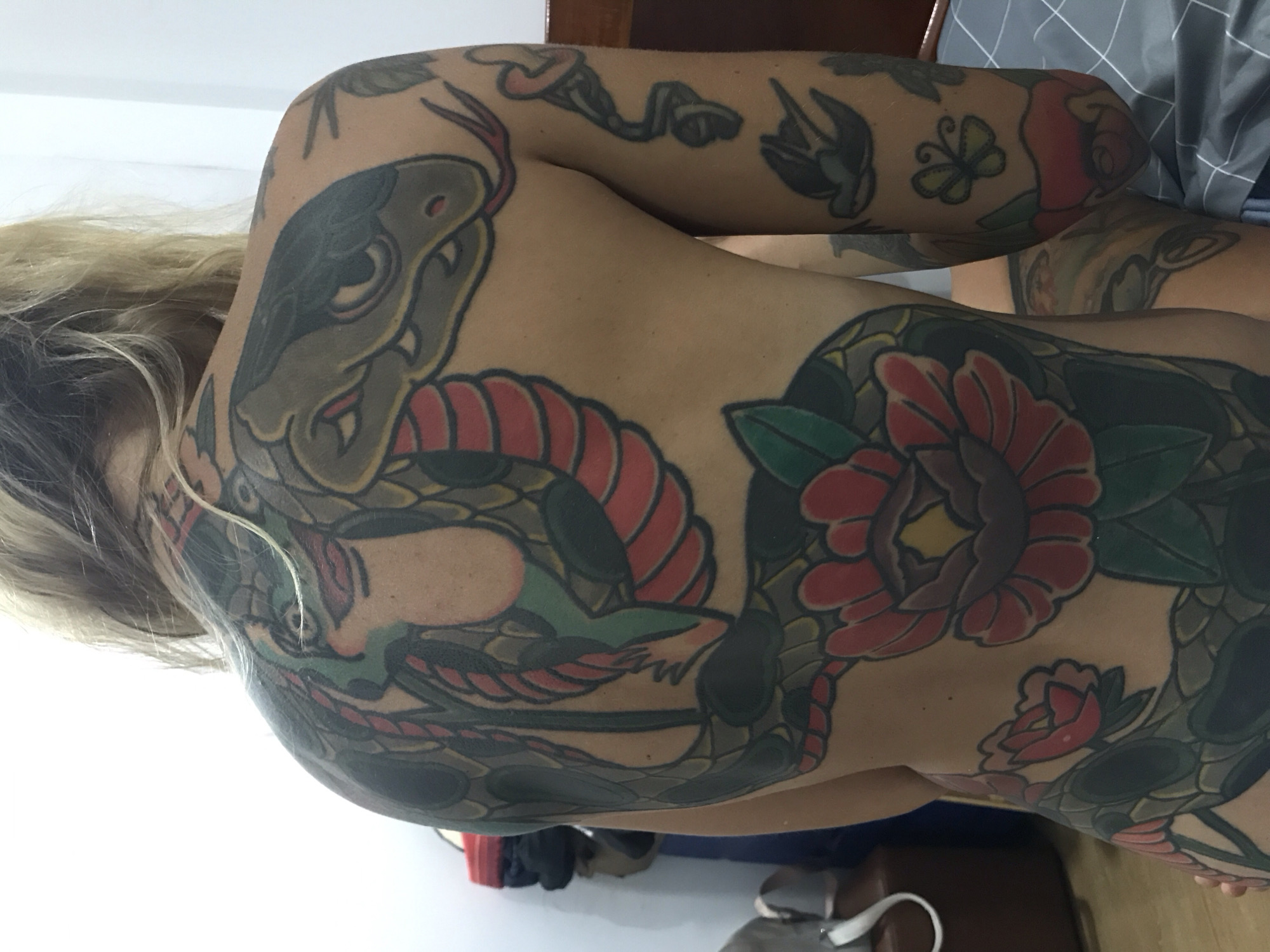 The image displays the back and right arm of a person, heavily covered with intricate tattoos. The person has long, wavy blonde hair that cascades down their upper back and shoulder.
The dominant feature is an extensive, colorful tattoo spanning the back and upper arm, rendered in a traditional or neo-traditional style. A large green and grey snake with distinctive red scales is depicted, coiling across the back. Its head, with dark eyes and fangs, is prominent on the left shoulder area. The snake is intertwined with several large, open red and pink flowers, possibly peonies, characterized by bold outlines and solid color fills. Green leaves and foliage complement the floral designs. Additional tattoos are visible on the right upper arm, including a dark swallow, a small yellow butterfly, and more floral patterns, consistent with the overall aesthetic.
The person's skin tone is light. The background is softly blurred, indicating an indoor setting. On the right side, a dark wooden furniture piece, possibly a headboard, and a grey fabric with a checkered pattern, likely bedding, are partially visible. The left side of the image is brightly lit, suggesting natural light, perhaps from a window. No activity is explicitly taking place beyond posing to display the tattoos. No text is visible in the image. The overall scene highlights the detailed body art. The location context for this media is Mũi Né, Vietnam.