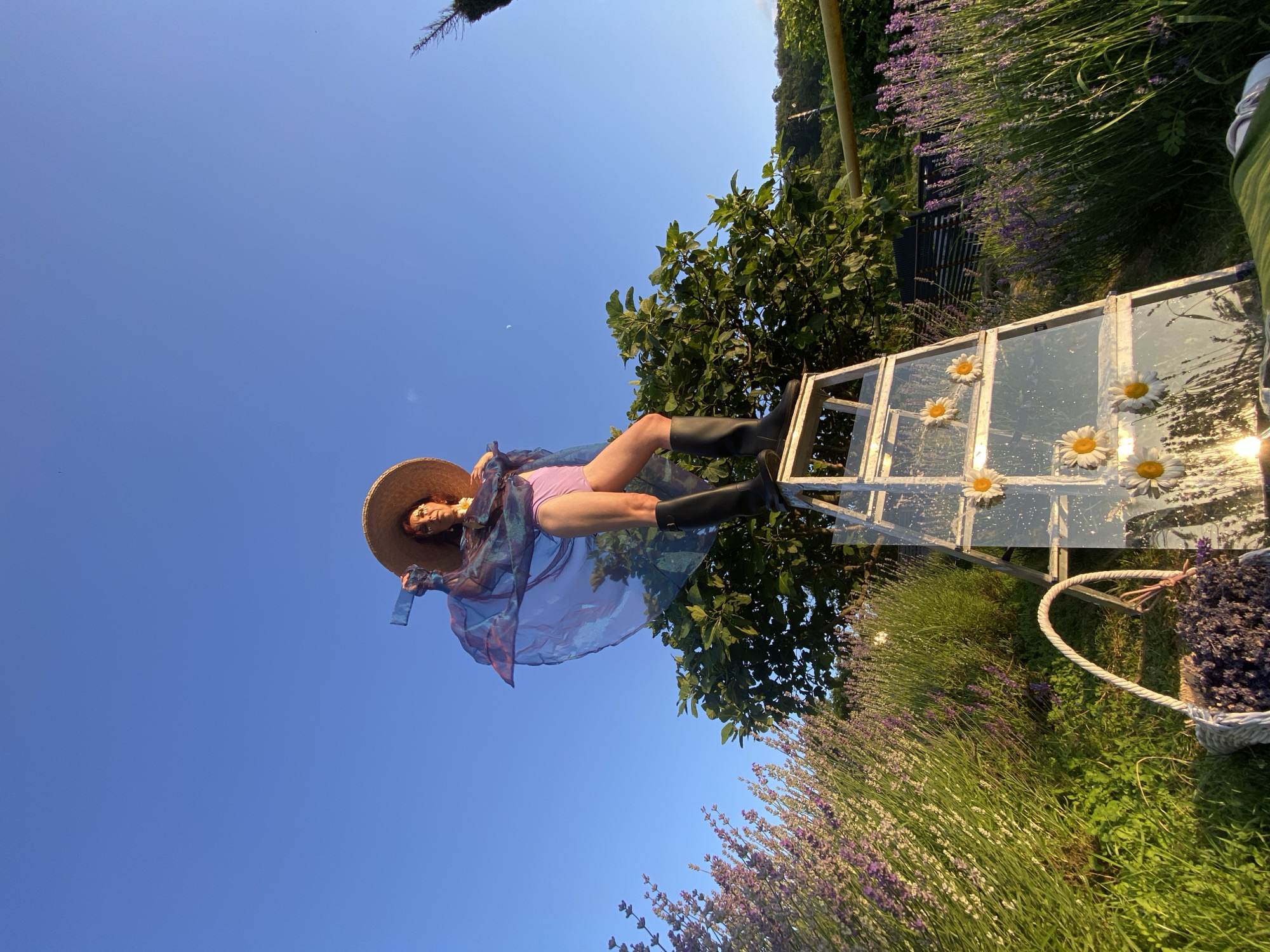 A person stands on a ladder decorated with daisies, in a field of lavender, under a clear blue sky. The person is wearing a straw hat, a pink swimsuit, a flowing sheer blue garment, and black boots. The ladder is leaning against a tree. A basket filled with lavender is in the foreground. The scene is likely a garden or a field in Verkhneye Uchdere, Russia, given the presence of lavender and the clear, sunny sky, suggesting an outdoor, rural setting.