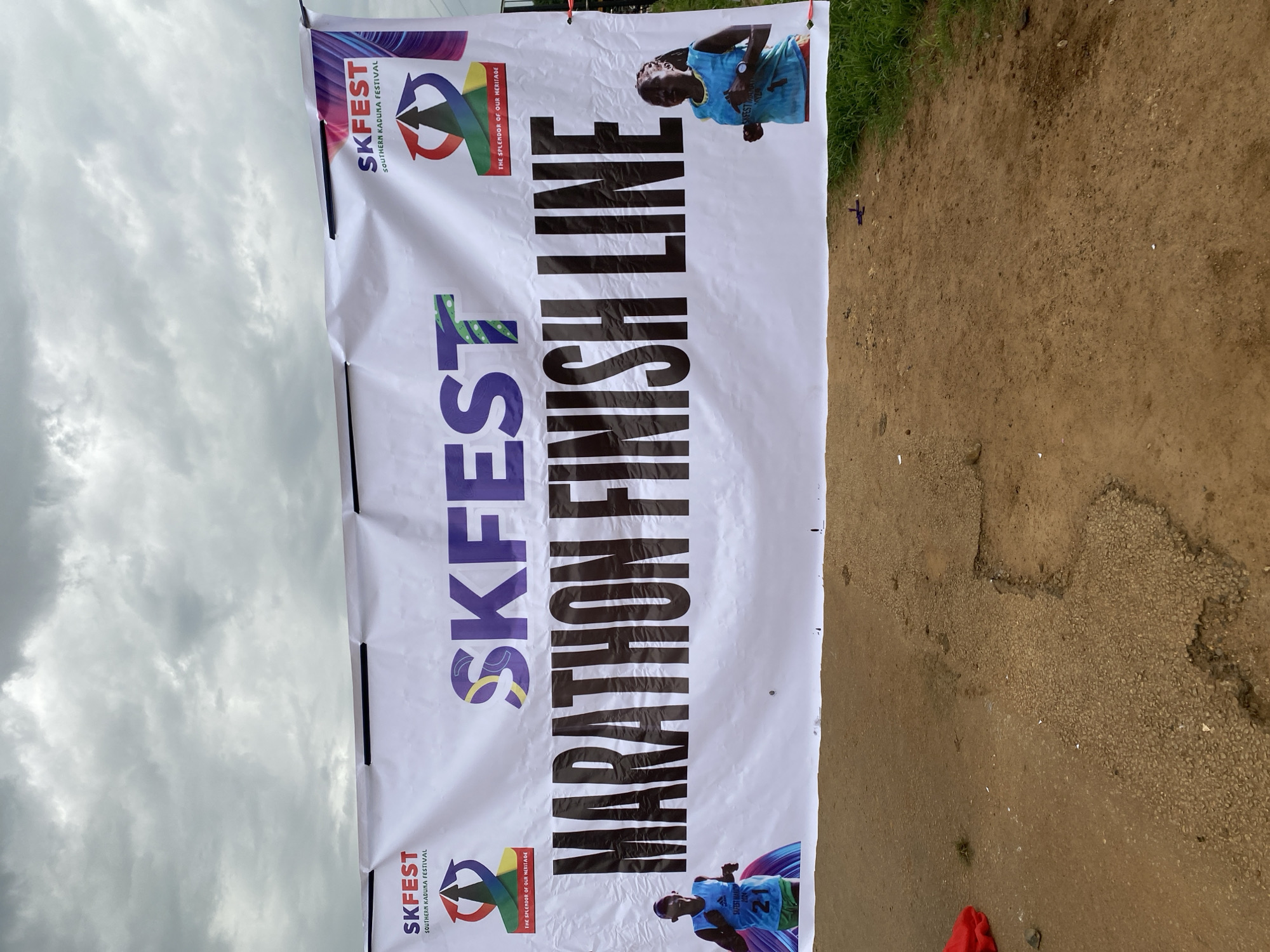A slightly rotated, medium-distance shot focuses on a white banner for the "SKFEST Southern Kaduna Festival" marking the "Marathon Finish Line." The banner is set against a backdrop of a cloudy sky above and a dirt ground below. The banner features the festival logo, including an arrow graphic and the phrase "THE SPLENDOR OF OUR HERITAGE." Two images of a runner are placed at the top and bottom of the banner. The runner in both images is wearing a blue jersey with the number "21" visible on one of the images. The bottom right corner of the image shows a red garment on the ground. The image appears to have been taken during the day in Maigizo, Nigeria.