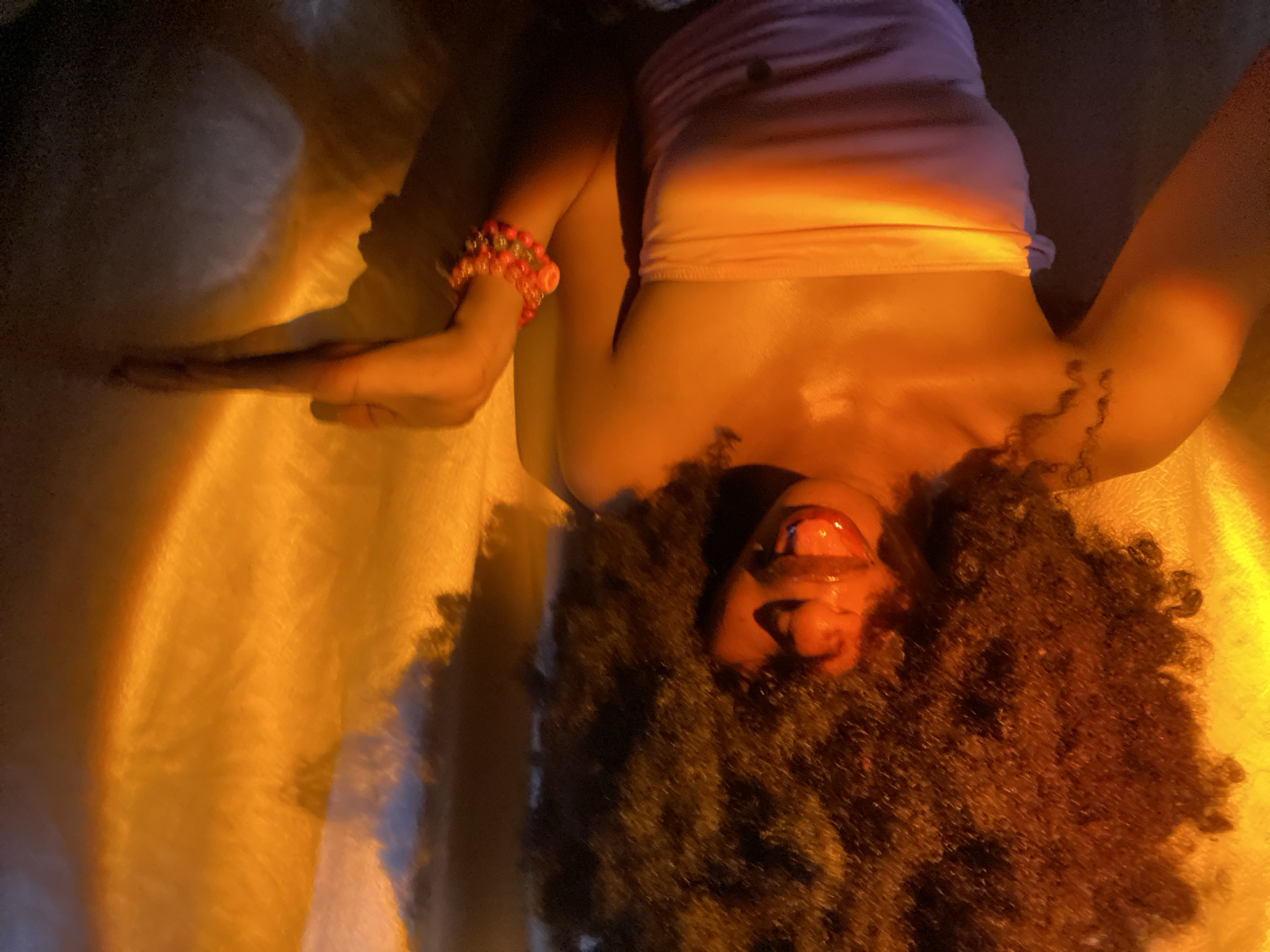 A person with brown curly hair is lying upside down. They are wearing a light pink tube top and have a beaded bracelet on their left wrist. The lighting is predominantly orange and yellow, with some rainbow effects on the torso. The background appears to be a textured, light-colored surface, possibly fabric or a wall with a textured finish. The city of Iranla, Nigeria, is not discernible in the image.