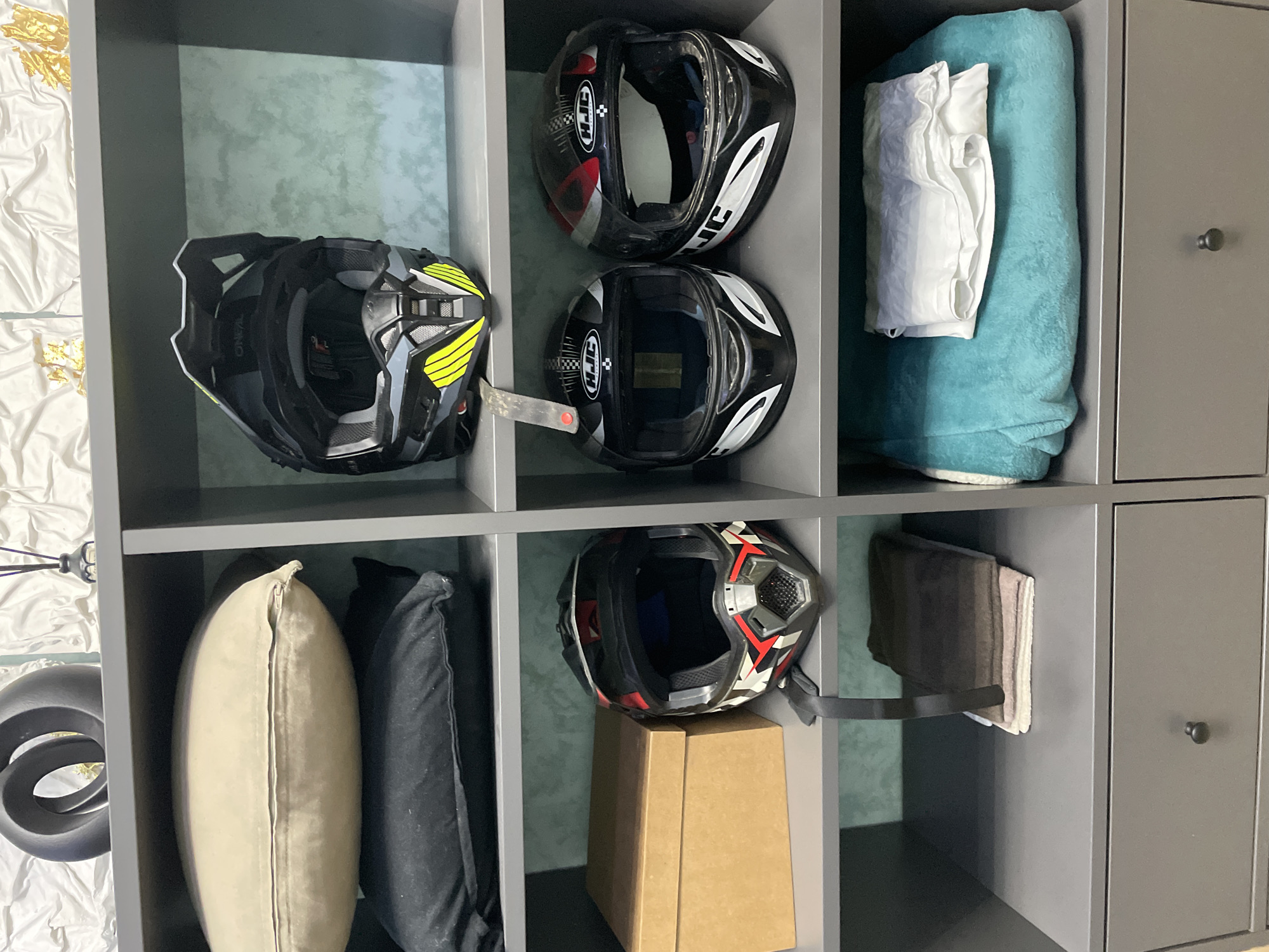 The image depicts a gray shelving unit or closet containing various items. On the top left shelf, there is a black motocross helmet with yellow accents. Next to it, on the top middle shelf, there are two black motorcycle helmets with white and red details. The top right shelf contains a folded light blue towel and a white folded shirt or cloth. The bottom left shelf has a beige pillow and a black pillow. Below the middle shelf, there is a black motorcycle helmet with red and white accents sitting atop a brown cardboard box. On the bottom right shelf, there is a folded brown towel. The shelves are part of a modular storage system with a door on the rightmost section. There are no people visible in the image. The location, Лесной Пейзаж Кп (шарапово), Russia, is not visually represented in the image.