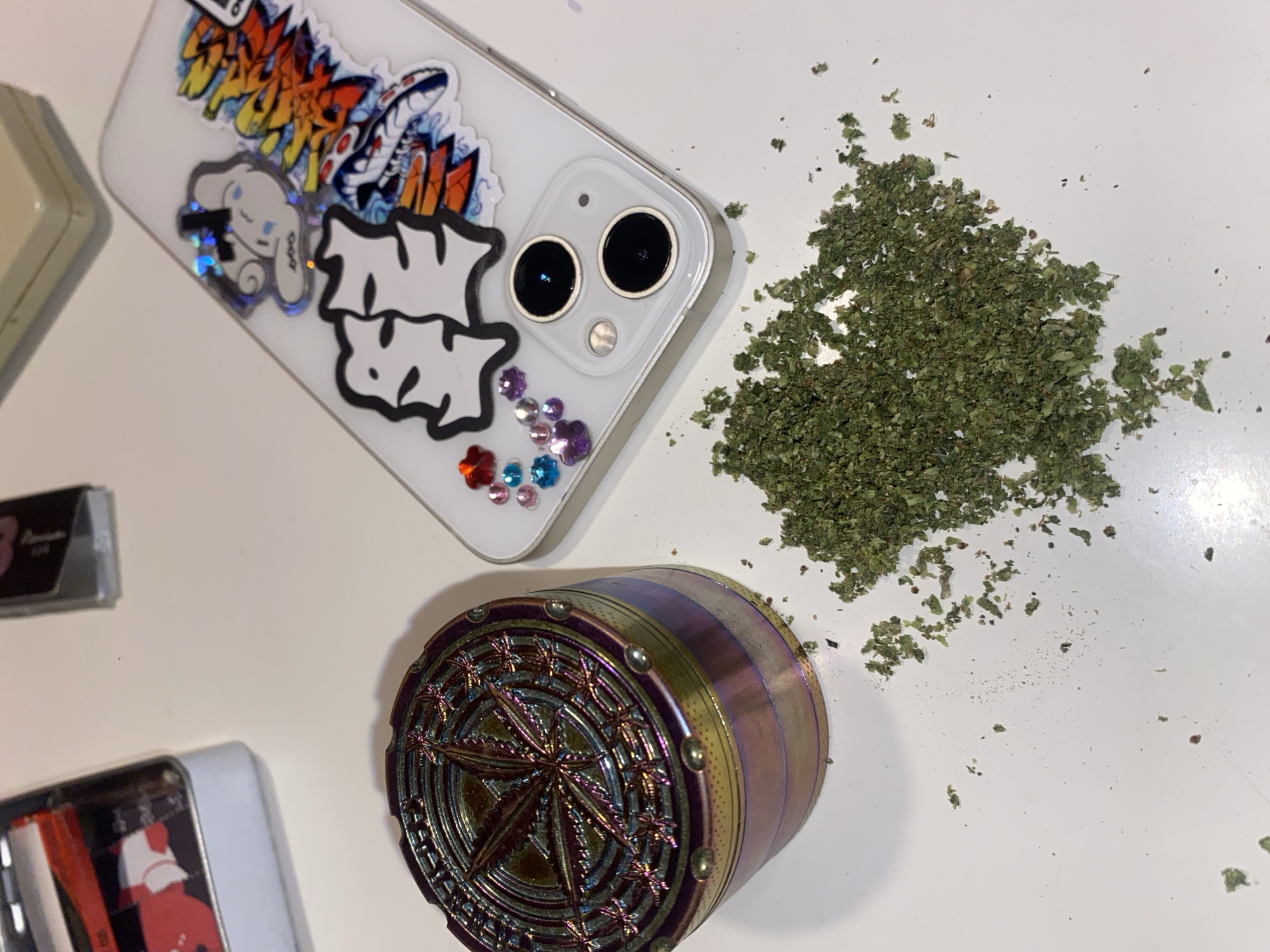 An indoor scene displays a collection of items on a smooth, light-colored surface, possibly a table or desk. A multi-colored, iridescent metal cannabis grinder, featuring a prominent cannabis leaf design on its lid, rests on the left side of the frame. To its immediate right and slightly below, a pile of finely ground, dry green plant material, consistent with processed cannabis, is scattered on the surface. A white smartphone, fitted with a clear protective case adorned with several decorative stickers and colorful rhinestones/gems, lies diagonally to the right of the grinder and above the plant material. The phone's dual camera lenses are visible at the bottom of its rear. Partially visible in the upper left corner is an open metallic tin containing red and white items. In the upper central background, a dark, rectangular folded object is discernible, while a light cream-colored object is partially visible in the upper right background. No individuals are present in the image, and no active interactions are depicted. The specific location within Buenos Aires, Argentina, cannot be determined from the visual evidence provided.
