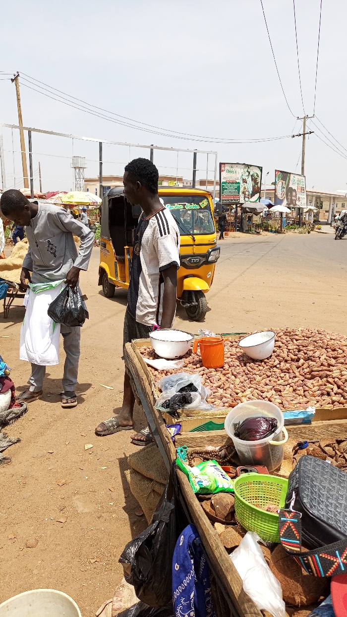 The image depicts an outdoor market scene in Barnawa, Nigeria, under bright daylight. In the immediate foreground, a makeshift wooden stall is laden with various goods. A large quantity of light brown, elongated produce, consistent with dried dates or groundnuts, occupies the main surface. Alongside these, white enamel bowls, an orange plastic cup, several clear plastic bags containing dark items, a green plastic basket, and a clear bucket holding a dark red cabbage are arranged. Other items on the stall include coconut halves, a patterned shoulder bag, a blue bandana, brown beads, and a small packaged item.
Two adult males are prominent in the foreground. On the left, a man wearing a light grey tunic with black embroidery, grey trousers, and brown sandals is partially bent over. He holds a black plastic bag in his right hand and appears to be adjusting a white cloth around his waist with his left hand. To his right, another man stands upright, wearing a white and black striped short-sleeved t-shirt, dark trousers, and dark patterned slide sandals. He faces left, away from the viewer.
Behind the individuals and the stall, a paved road is visible. A yellow TVS-branded auto-rickshaw (Keke Napep) with "Garden Hukba KFC" text is positioned on the road, facing left. The surrounding environment includes dry, light brown earth transitioning to the asphalt road. In the midground and background, utility poles with overhead wires are present. Multiple billboards are visible, including one advertising "Pukka's" and another with "Garden Hukba KFC" branding. Various buildings, distant market stalls with umbrellas, and a few individuals, including a person on a motorcycle, are discernible further down the road. The sky is clear and light-colored, indicating a sunny condition.