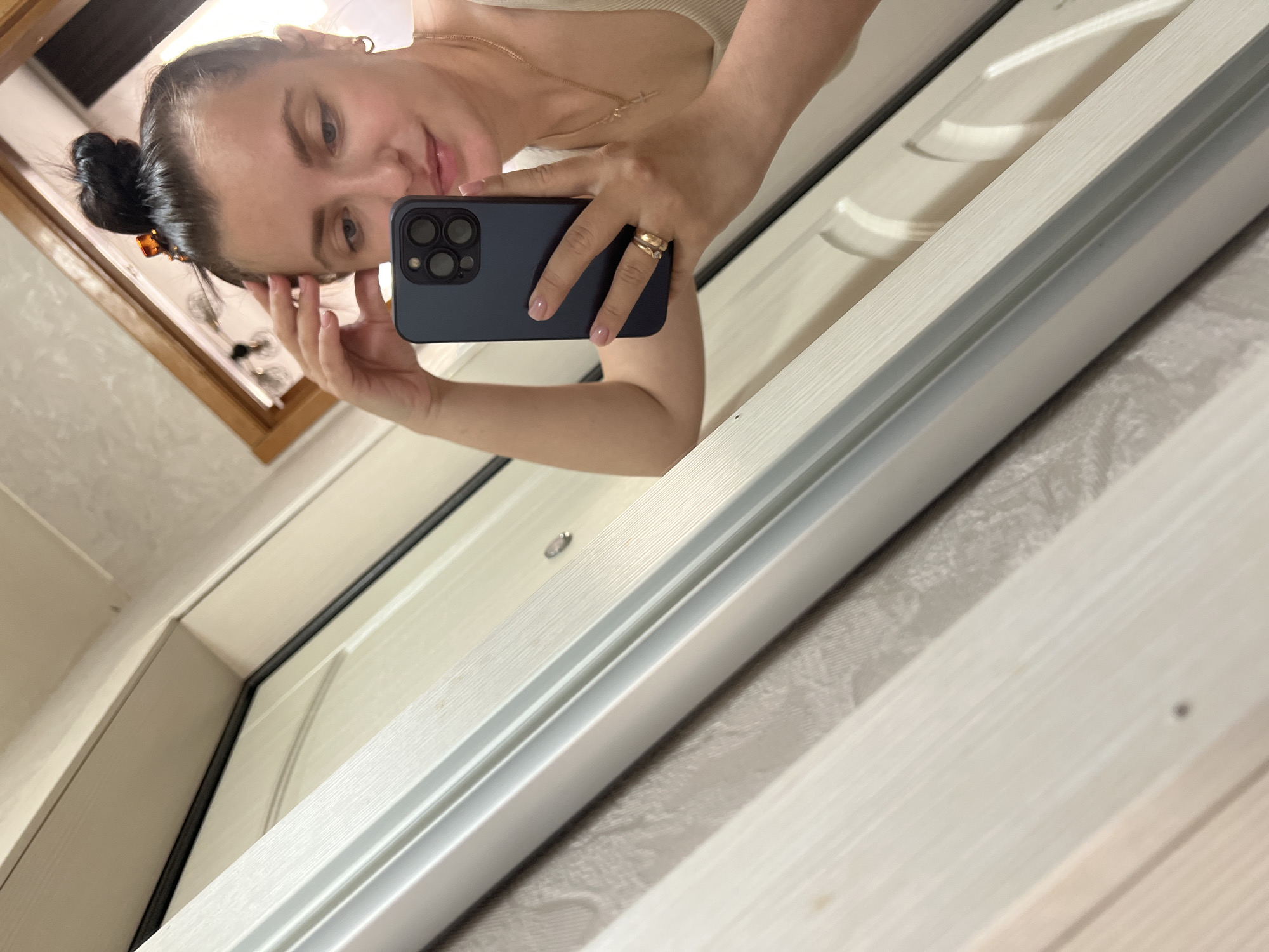 This media file is a portrait of a woman taking a selfie in a mirror. The woman has dark hair pulled back in a bun and is wearing a light-colored top and a necklace. She is also wearing two rings on her right hand. The background appears to be a room with white doors and textured walls, suggesting an indoor setting. The lighting is bright, indicating it could be daytime. The woman's expression is a slight smile, and she is touching her forehead with her left hand. The photo is taken from a low angle, and the camera is held in her right hand. There is no visible text in the image.