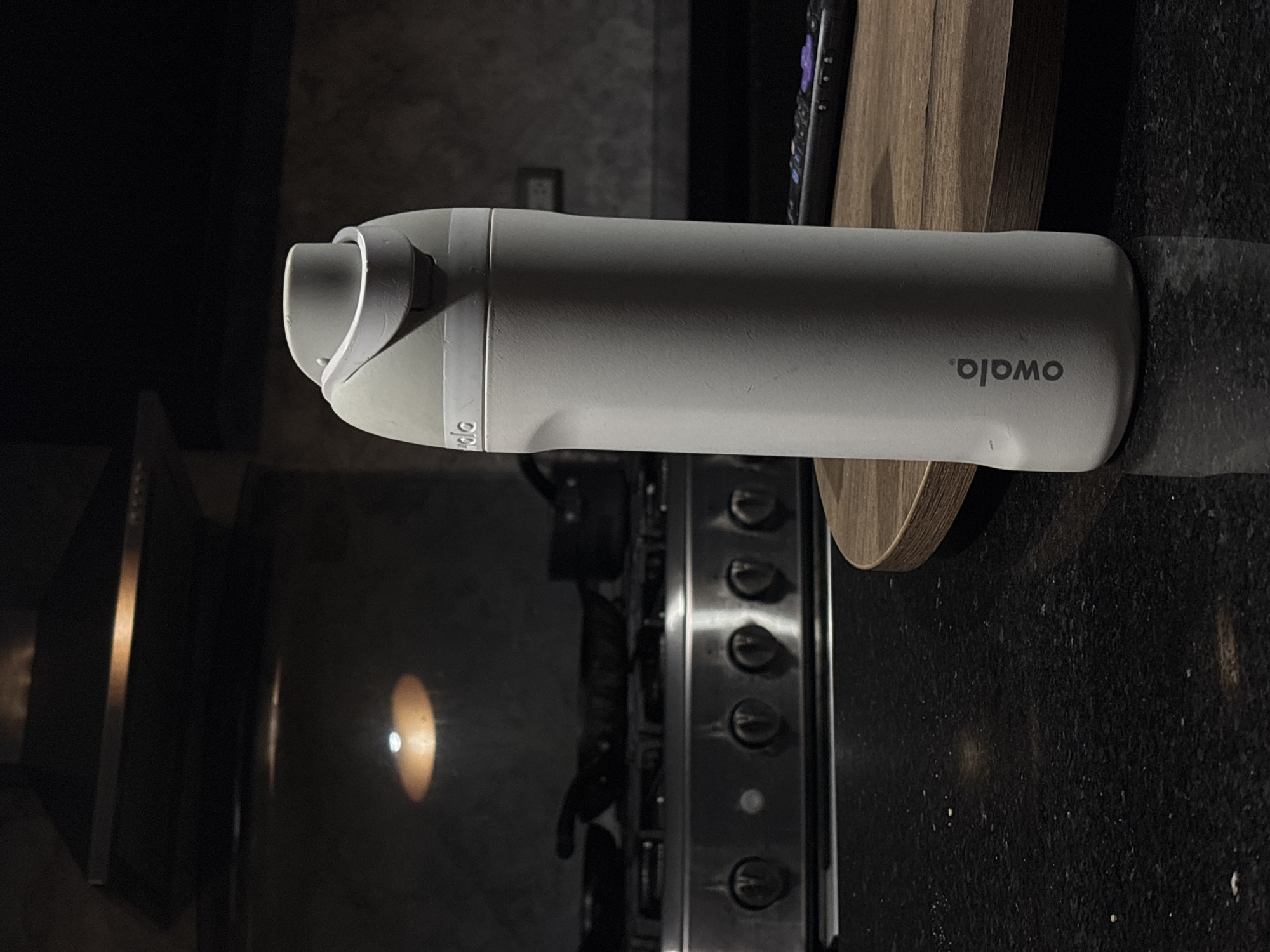 The image shows a gray Owala water bottle lying on a black countertop in a kitchen setting. The bottle is oriented horizontally, with the word "Owala." visible on its side. Behind the bottle is a wood cutting board, a black remote control, and a stainless steel gas stove with multiple control knobs. To the left of the stove, there is a glimpse of a black oven door and cabinet. A light reflection can be seen on the oven door. The overall lighting in the scene is dim. This image does not show the city of Santa Clara Ocoyucan, Mexico.