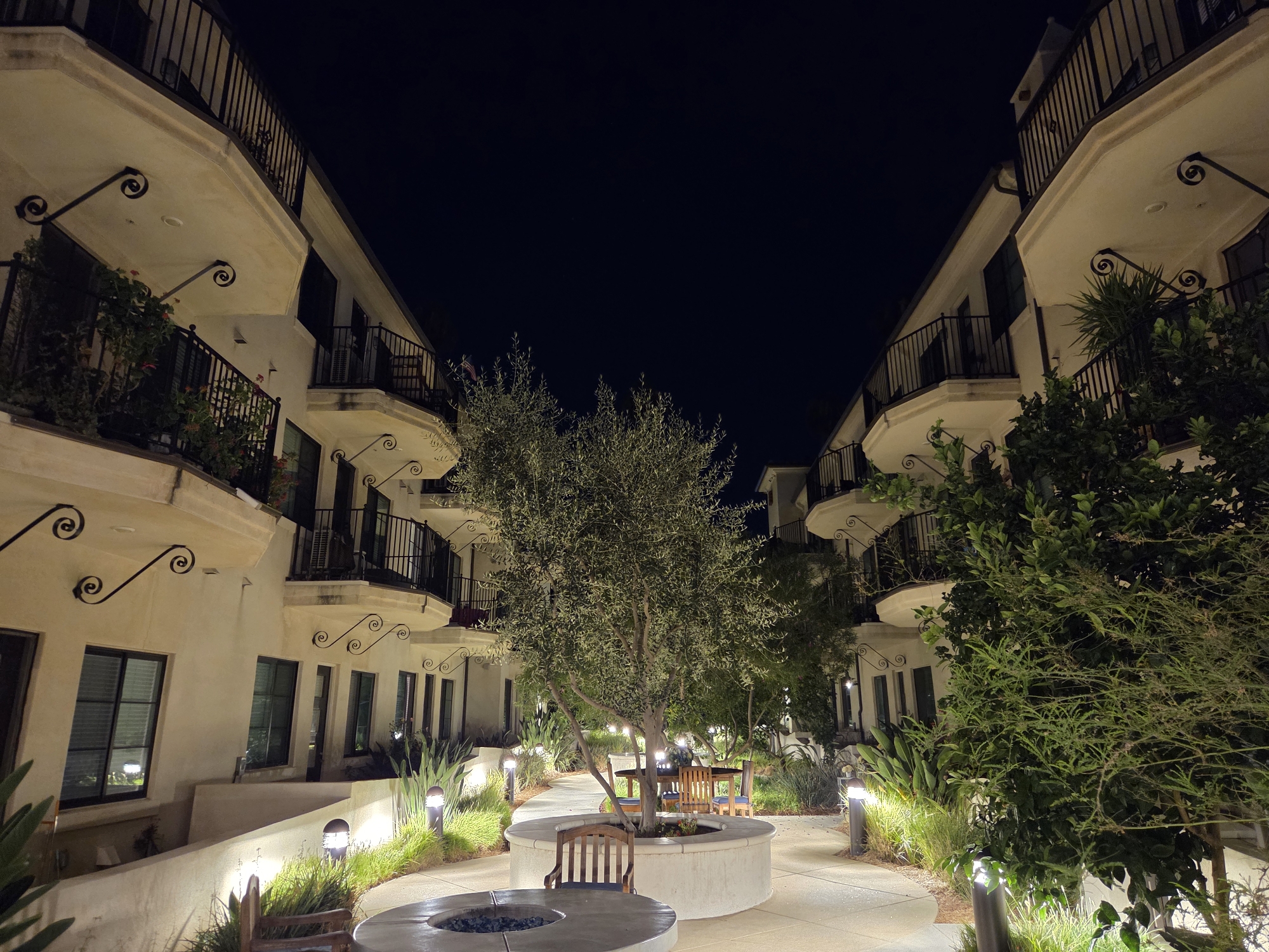 The image shows a courtyard area at night, likely in an apartment complex or hotel, located in Glendale, United States of America. The scene is illuminated by artificial lighting. On both sides of the courtyard are multi-story buildings with cream-colored facades and black wrought-iron railings on balconies and walkways. The buildings appear to be residential or hotel accommodations.

In the center of the courtyard, a pathway winds through landscaped areas featuring various plants and grasses. A prominent circular planter contains a medium-sized tree, possibly an olive tree, with its branches illuminated by overhead lights. Around the planter, there are wooden chairs and tables. In the foreground, a circular fire pit is visible on a paved surface. The overall atmosphere is calm and serene, with the dark night sky providing a stark contrast to the lit architecture and greenery. No people are visible in the image.