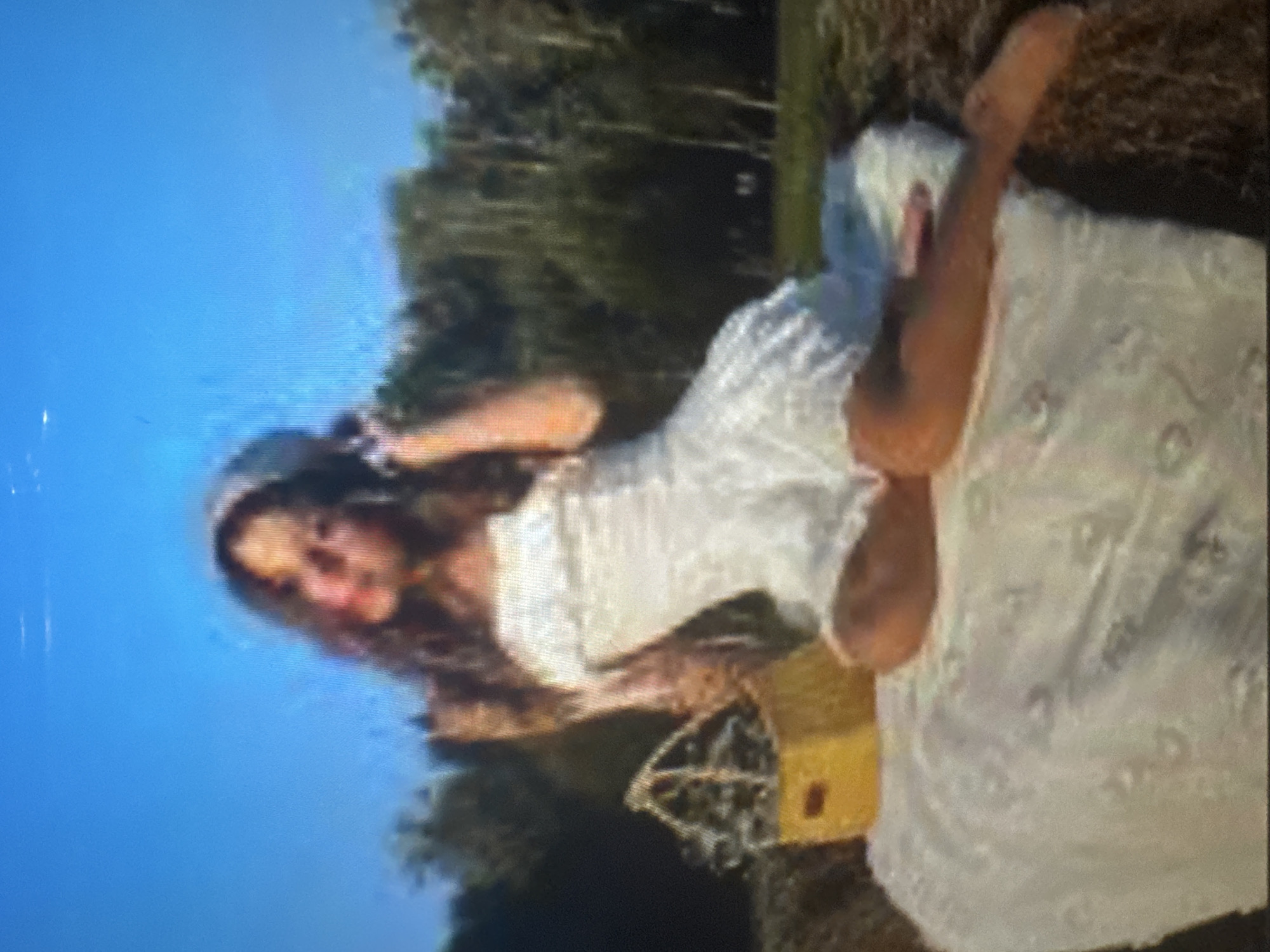 The image, when viewed in its correct upright orientation, captures a young woman posing outdoors on a bright, clear day. She has long, wavy dark hair and is wearing a light-colored, possibly white, textured dress or top. A hat or headband is visible on her head, with one hand placed near it. She is lying on her side on a surface that appears to be dry grass or hay, with a white patterned blanket or cloth spread beneath her. Her legs are bent, and one leg is raised, exposing the sole of her foot. A yellow woven basket with a handle is positioned in front of her. The background consists of a clear blue sky on one side and dense green trees or foliage on the other, indicating a natural, open-air setting like a field or park. The image quality is low, appearing blurry and pixelated with visible horizontal lines, suggesting it might be a photograph of a screen. No discernible text is present. The scene conveys a relaxed and serene atmosphere, possibly a moment from a leisurely outing or a photo session.