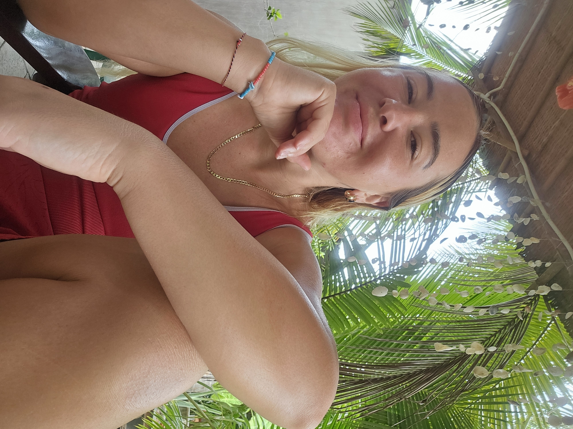 A young woman with blonde hair and light skin is pictured from the chest up, wearing a red tank top with a gray trim. She is seated outdoors, with her left arm bent and resting on her crossed legs. Her right hand is placed under her chin, and she is looking towards the camera with a slight smile. She is wearing a gold chain necklace, gold hoop earrings, and a bracelet with blue and red beads on her right wrist, and a thin red string bracelet on her left wrist. Behind her, palm fronds and a thatched roof are visible. The lighting suggests it is daytime. The location is not specified within the image itself.