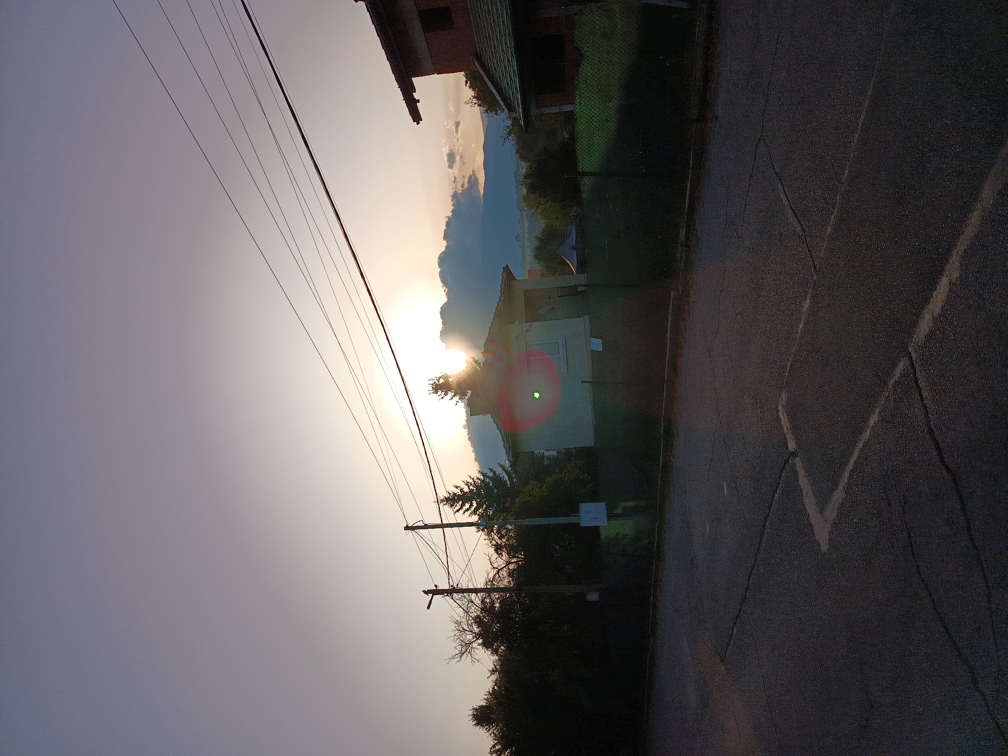 The image shows a sunset scene in Kralev Dol, Bulgaria. The sun is low in the sky, partially obscured by clouds, casting a warm glow. Power lines stretch across the frame, silhouetted against the sky. In the foreground, a paved road with visible cracks and painted lines suggests a street or parking area. Behind the road, a chain-link fence separates it from a property with a white house and a red-tiled roof house. Several trees, including a prominent evergreen, are scattered around the houses. The overall atmosphere is peaceful and rural.