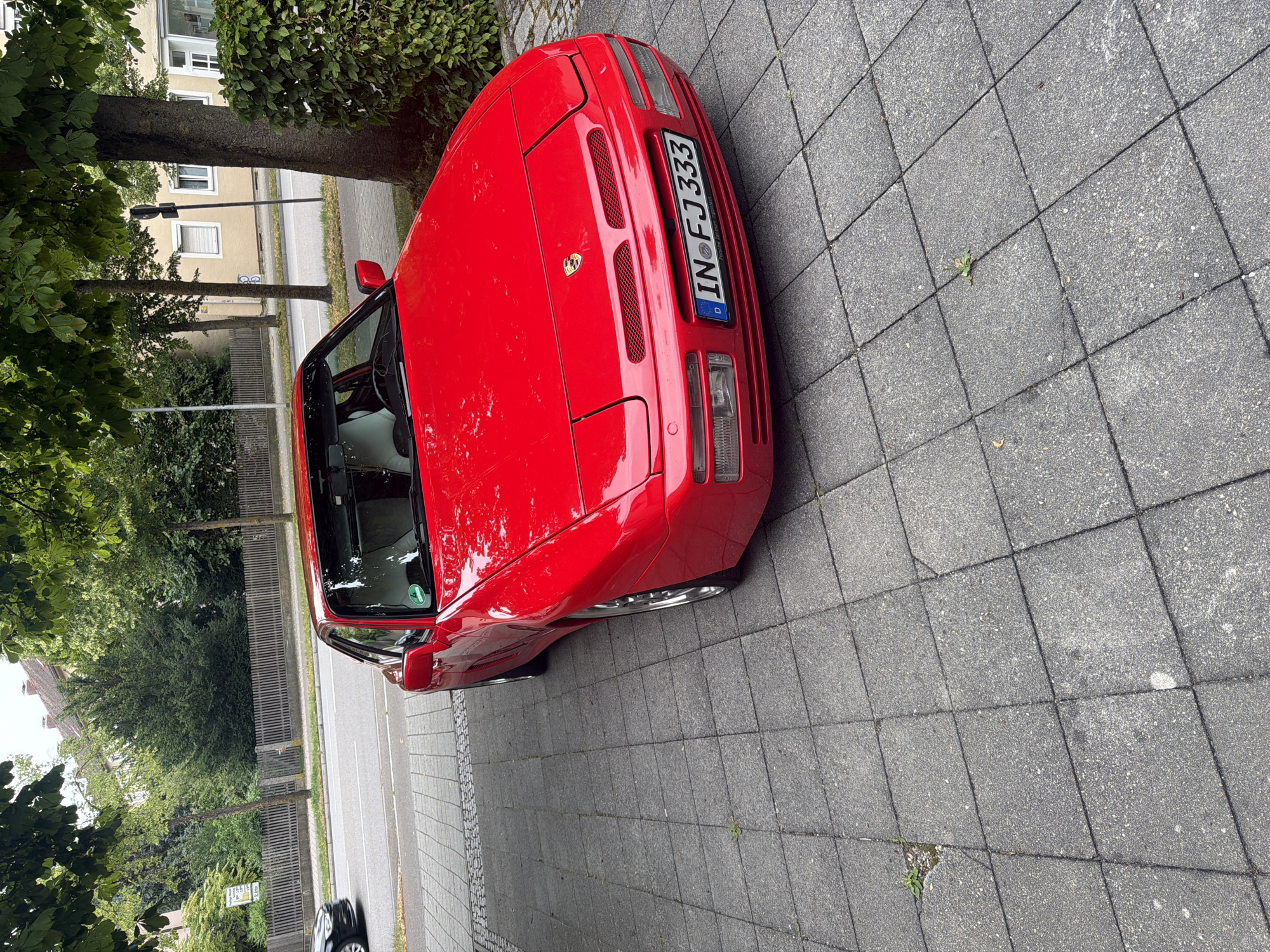 A bright red Porsche 944 is parked on a grey paver driveway in front of a building in Ingolstadt, Germany. The car is angled towards the viewer, with its front bumper and hood prominently displayed. The license plate reads "IN FJ 333". The car is under the shade of a large tree with green leaves. In the background, a street with a black car is visible, as are buildings and more trees. The driveway is made of square concrete pavers with visible cracks and some weeds growing in them.