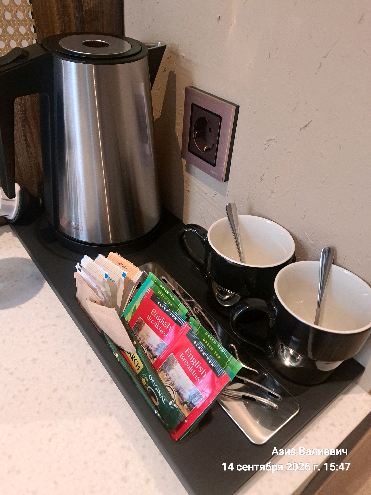 An indoor, eye-level, medium shot captures a hospitality setup on a light-colored countertop. A stainless steel electric kettle is prominent on the left. To its right, a black tray holds two black mugs, each containing a silver spoon. In front of the mugs, a small basket-like holder contains several tea packets and what appear to be sugar or creamer sachets. Some of the visible tea packets are labeled "English Breakfast" and "Green Tea." A square, light-brown electrical outlet with a circular socket is mounted on a textured, light-colored wall behind the setup. The scene is illuminated by ambient lighting, casting soft shadows. The bottom right corner of the image includes text in Russian that reads "Азиз Валиевич 14 сентября 2026 г. 15:47", indicating a name and date/time stamp. This scene is likely in a hotel room or guesthouse in Tashkent, Uzbekistan, given the Russian text and the common amenities provided.