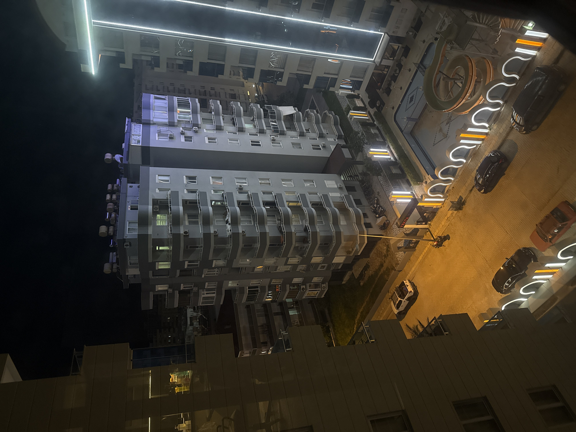 The image depicts a nighttime aerial view of a city, likely Unknown, Unknown, showing multiple multi-story buildings. The buildings appear to be residential or commercial apartments, characterized by rows of windows and balconies. Exterior lighting illuminates the buildings and adjacent streets, including ornamental lights along the pathways. Vehicles are visible on the street, indicating vehicular activity. A building under construction is noted in the foreground.