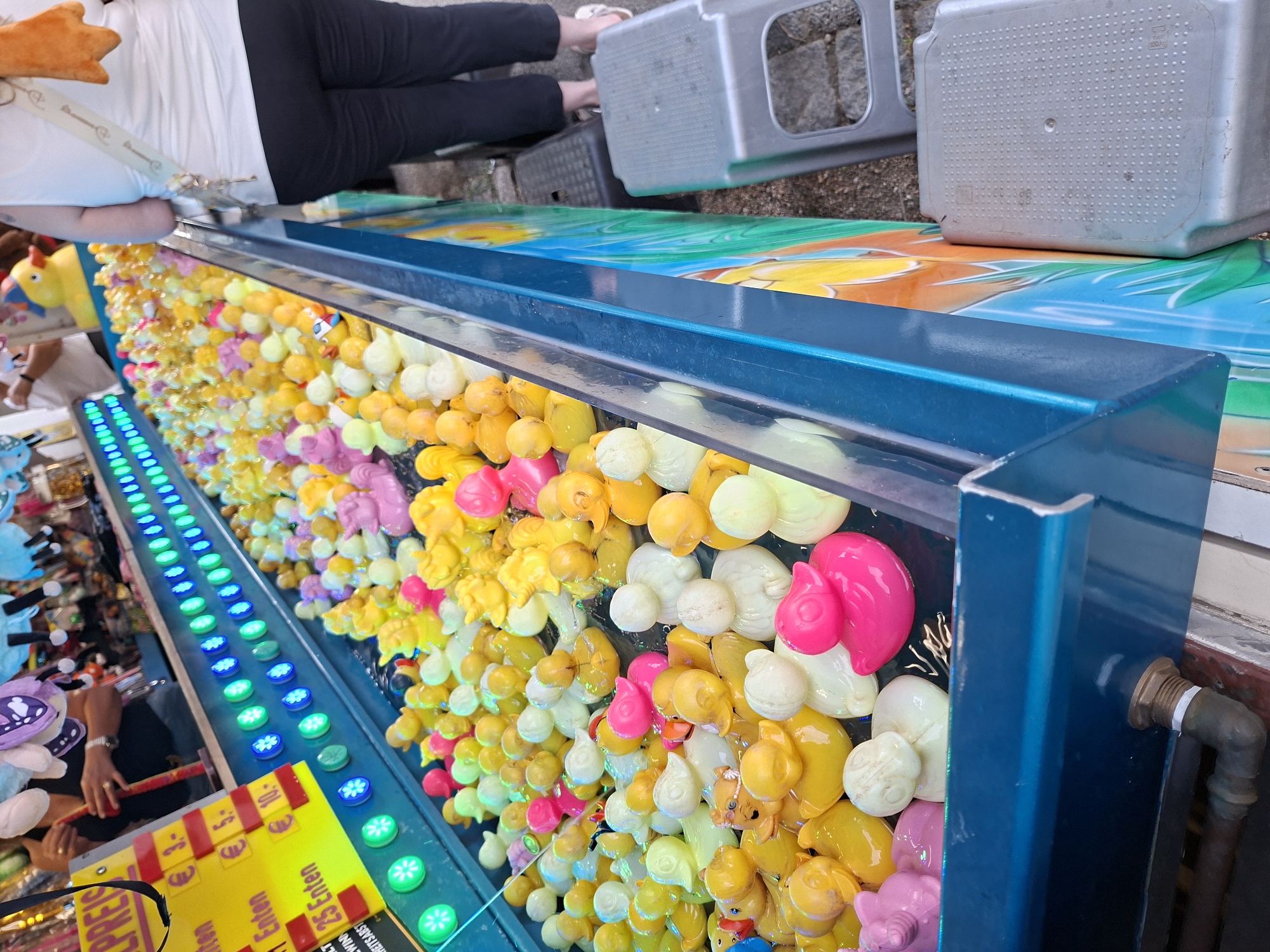 This media file shows a close-up, slightly angled view of a carnival game stall filled with numerous small, brightly colored rubber ducks. The ducks are predominantly yellow, with some pink and white ones mixed in. They are packed tightly together in a water-filled trough, suggesting a "hook-a-duck" style game.
In the background, the legs and lower torso of a person wearing black leggings and a white top are visible, sitting on a bench or stool, their feet clad in white sandals. This person appears to be operating or observing the game.
To the left of the ducks, a row of illuminated green and blue LED lights runs along the edge of the stall. Below the ducks, a yellow sign with some German text is partially visible. The text that can be read includes "PREIS", "10.", "Enten", and "25 Enten". This indicates prize information related to the number of ducks one might "catch" or win.
Further to the right, a large, grey, plastic container with a textured surface is visible, and beyond that, a glimpse of a stone-paved area. The overall setting appears to be an outdoor event, likely a fair or carnival, given the presence of the game stall and the casual attire of the person. The lighting suggests it could be daytime, though the specific time and weather conditions are not explicitly clear. The ducks themselves are the primary focus, presented in a vibrant and playful manner, indicative of a fun, possibly competitive, activity.