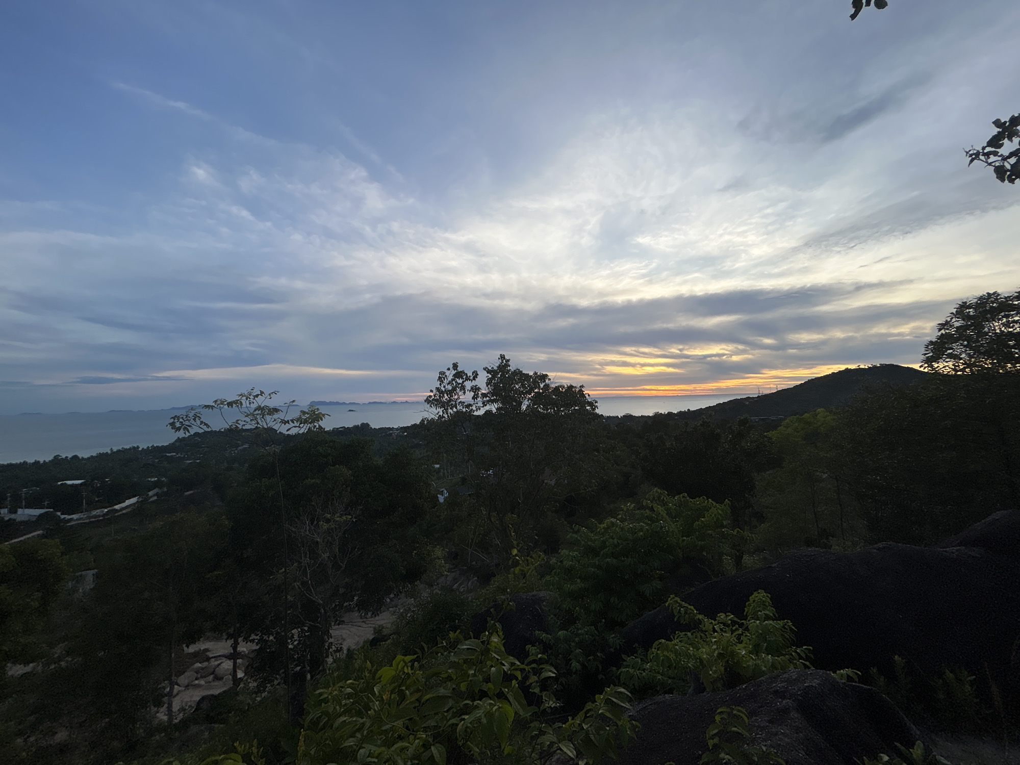 The image shows a landscape in Baan Sri Thanu, Thailand, captured at dusk. In the distance, a body of water is visible under a cloudy sky exhibiting sunset colors. The foreground features lush vegetation and rocky terrain. A hill is visible to the right of the water, and tree branches are in the upper right corner of the frame.