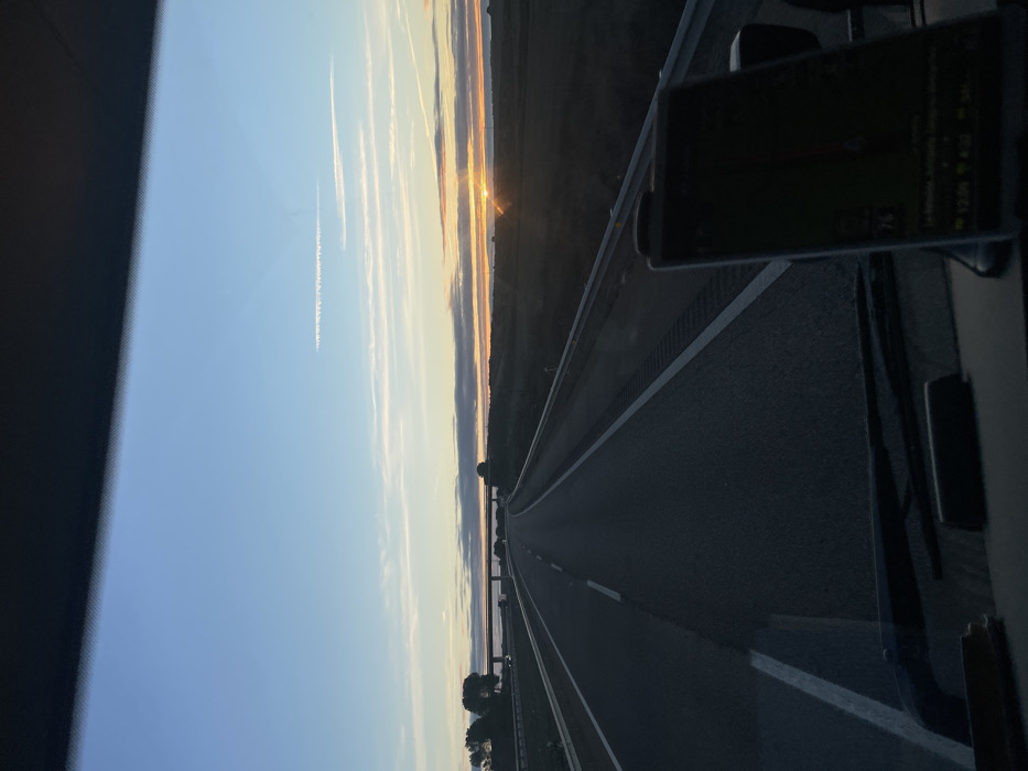 A highway stretches into the distance, illuminated by a setting sun. The road is bordered by fields and trees. Jet contrails mark the blue and orange sky. A navigation device is mounted on a dashboard to the right of the view, showing map details and the time. The photo is taken from the interior of a vehicle looking forward. The image is rotated.