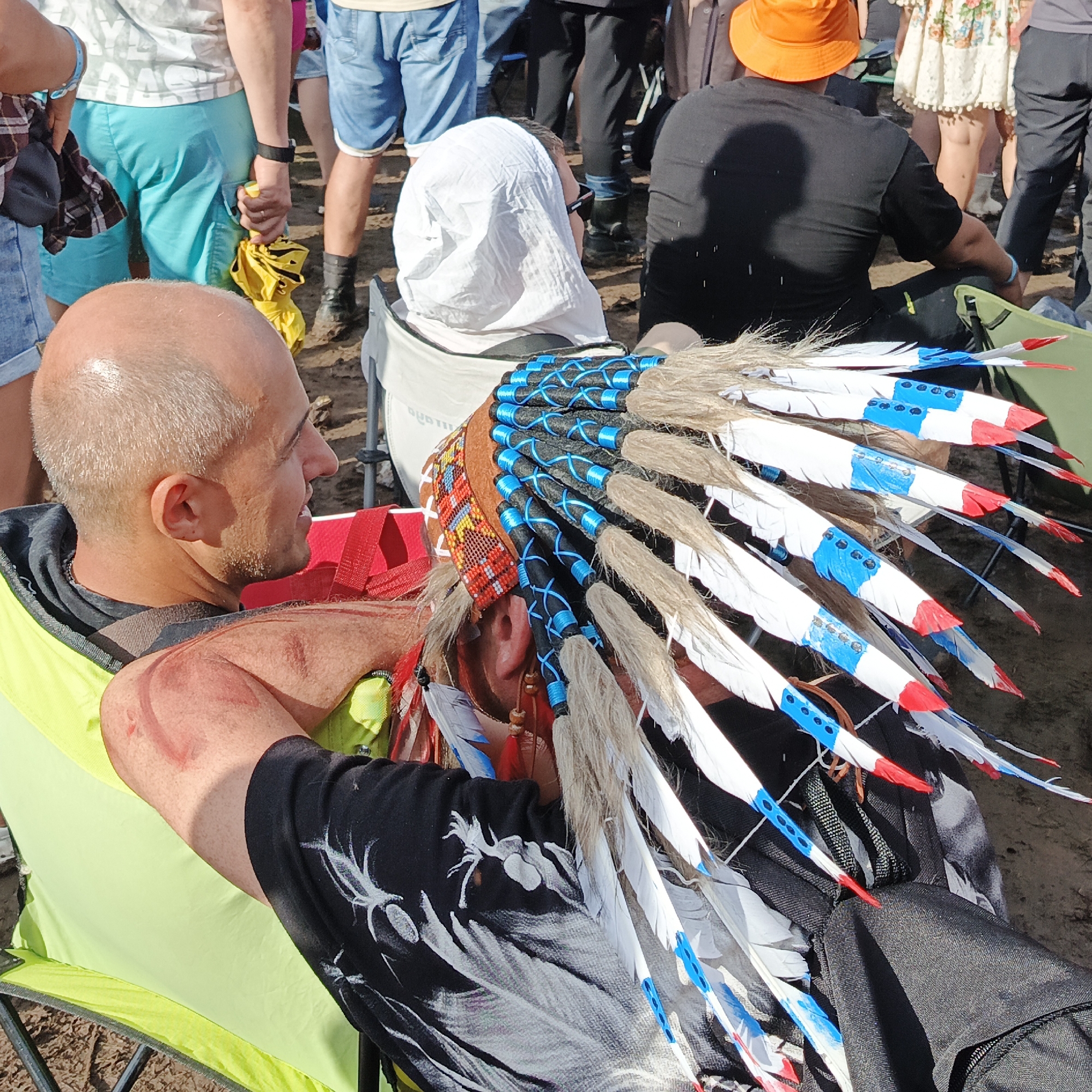 In the city of Lagunovo, Russia, a man wearing a Native American-style headdress with white, blue, and red feathers is seated in a lime-green camping chair. He has a shaved head and is wearing a black shirt with white avian-like graphics. The headdress is adorned with blue beads and intricate patterns. His right arm, resting on the armrest of the chair, shows a red mark. Another person is visible in the background, partially obscured, wearing a white head covering. Various other individuals and their legs, dressed in casual attire such as shorts and pants, are present in the background, suggesting an outdoor gathering or event. The ground appears to be dirt or sand.