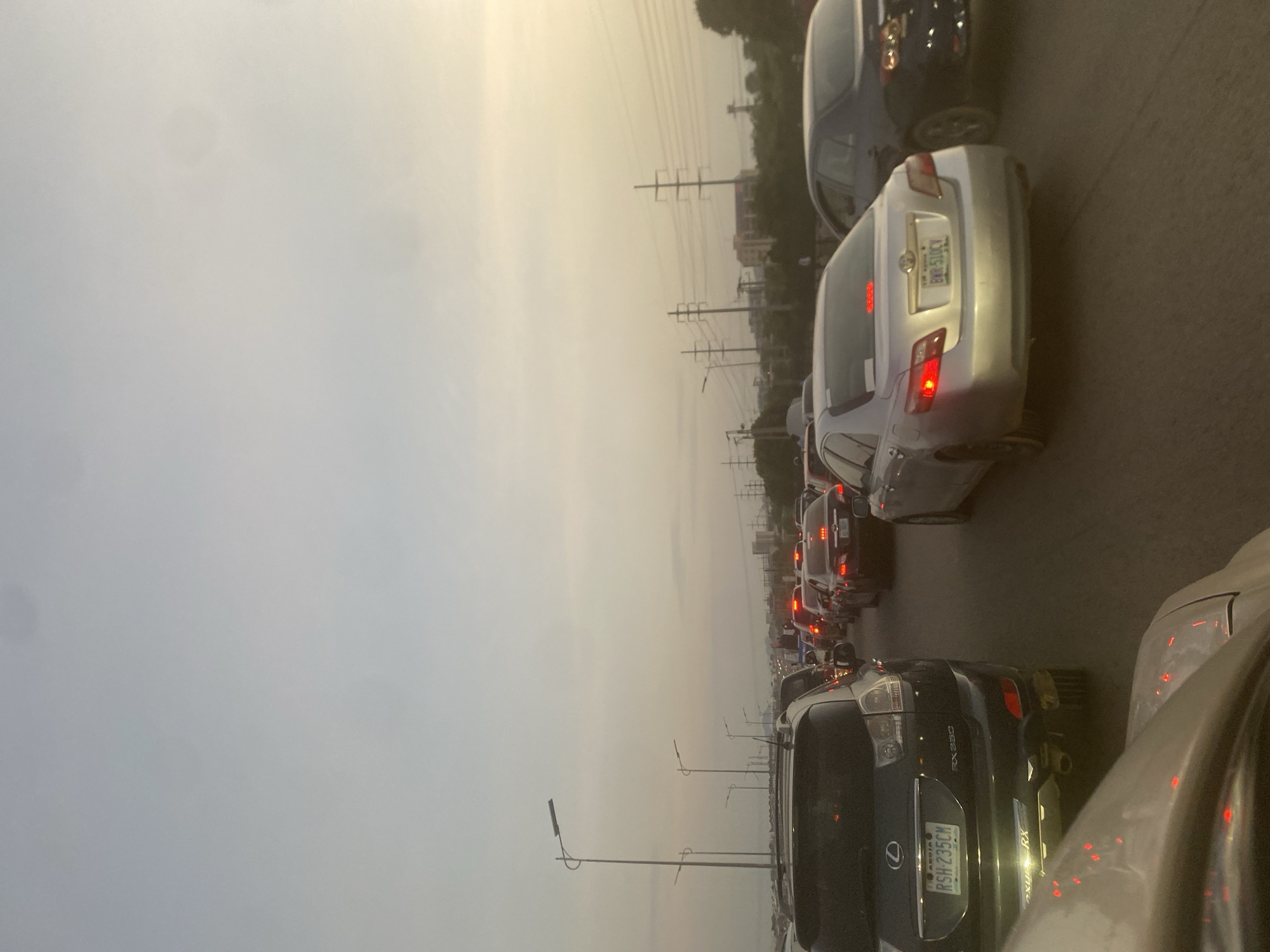 The image depicts a congested highway with multiple lanes of stationary vehicles during what appears to be rush hour. The perspective is from a vehicle looking forward, capturing a long line of cars, SUVs, and trucks. Overhead streetlights are visible along the side of the road, and in the distance, power lines and buildings can be seen. The sky is overcast, with the sun setting or rising, casting a warm, diffused light. The scene is characterized by the stillness of traffic, with brake lights illuminating the road ahead. There are no visible people outside of the vehicles. The location is identified as Kuchingoro, Nigeria.