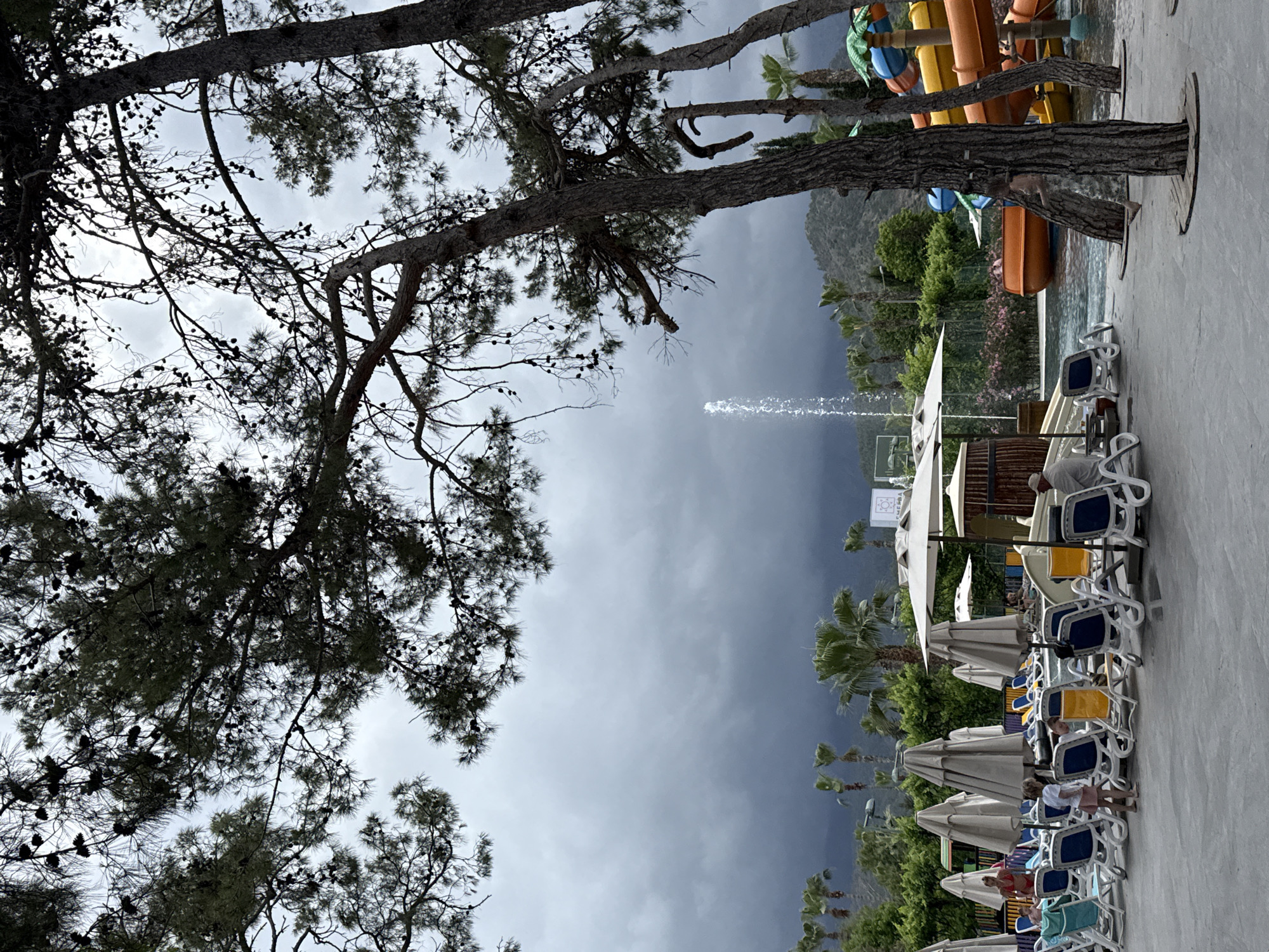 The image depicts an outdoor scene at a resort in Kemer, Turkey. Dark, cloudy skies hang over a mountainous backdrop. In the foreground, a mature pine tree with visible cones frames the upper left of the view. Moving towards the right, waterslides are present, adding a leisure element to the setting. Adjacent to these, a row of sun loungers with umbrellas occupies the right side of the image. People are situated under some of the umbrellas. A fountain shoots water upwards in the center of the frame.