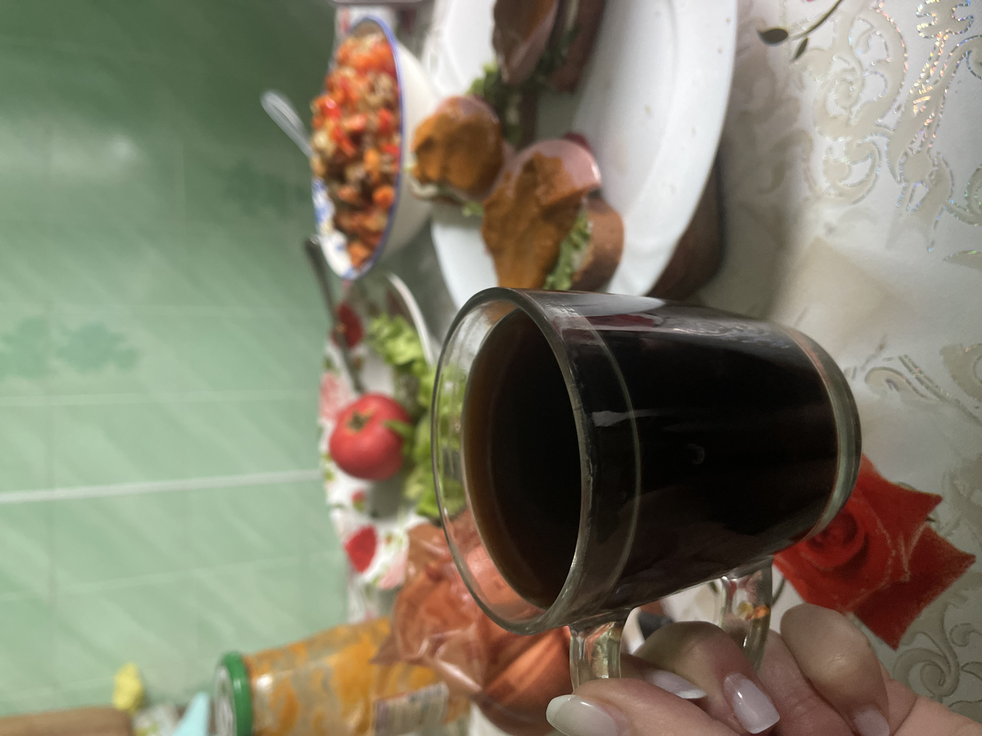 A close-up shot captures a clear glass mug filled with dark coffee held by a person's hand with light-colored nails. In the background, a table is laden with food items. To the left, a glass jar with an orange substance and a transparent plastic bag containing a few items are visible. Further back, a plate with a red tomato and green lettuce is present, along with a bowl of salad. To the right, a white plate holds several open-faced sandwiches topped with what appears to be meat and sauce, with one sandwich in focus. The setting suggests a meal in a kitchen or dining area, likely within a home in Красное, Russia. The overall atmosphere is casual and domestic.