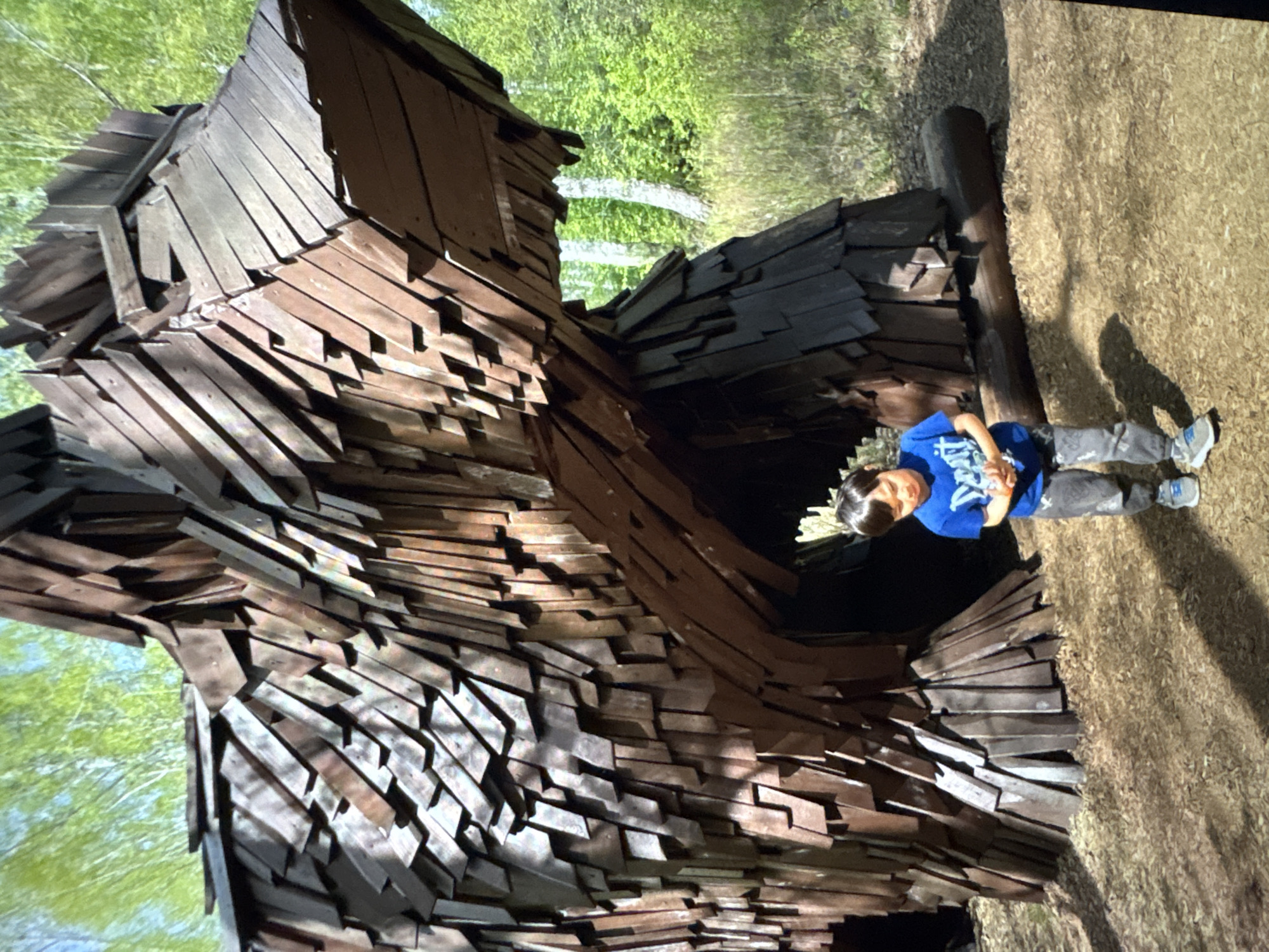 A young boy wearing a blue shirt and gray pants stands on a dirt-covered ground near a large wooden structure made of many overlapping pieces of wood. The structure has an opening or entrance where the boy appears to be looking into. The background shows trees and greenery. The location is Ембаево, Russia.