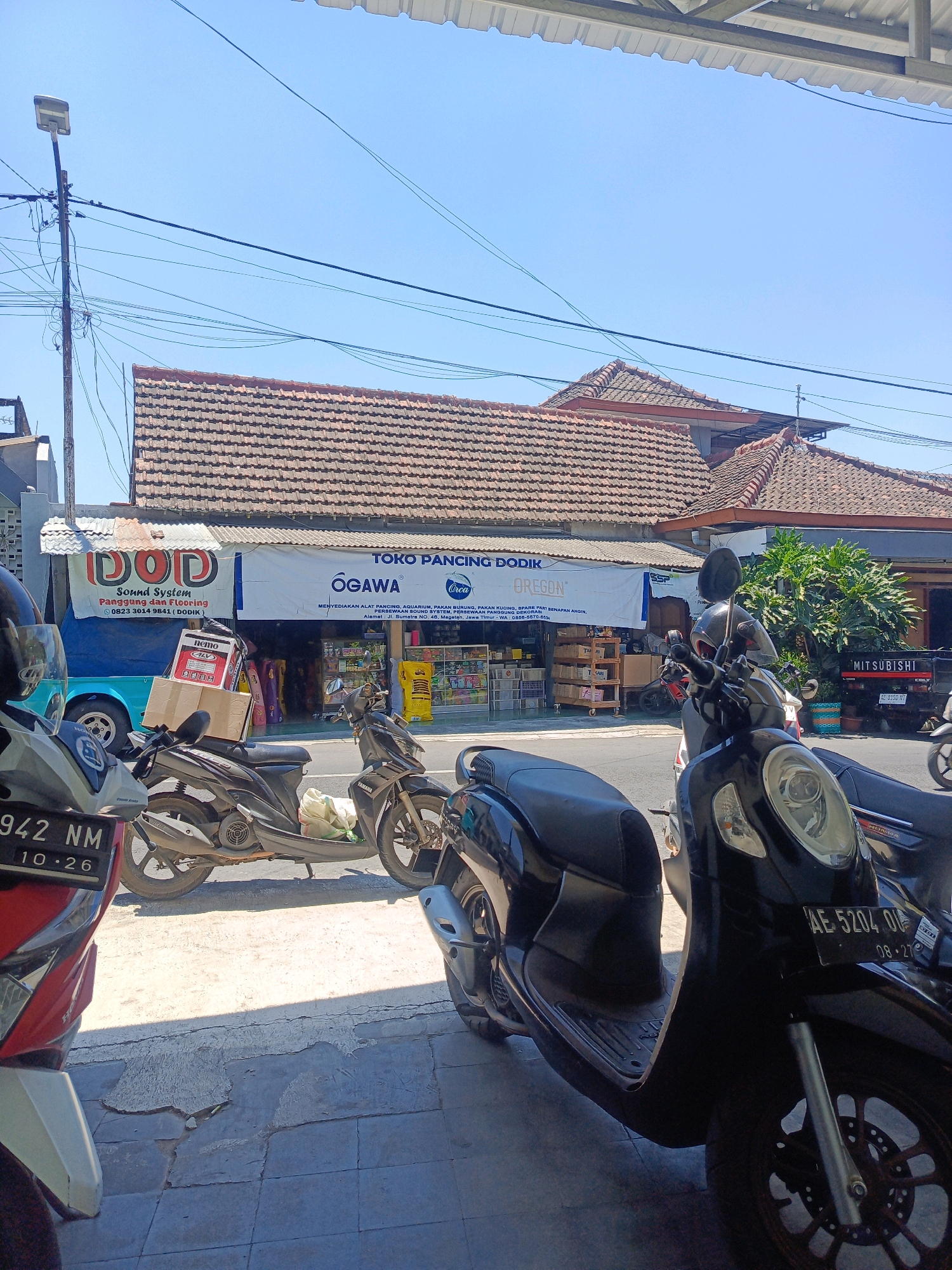 The image captures a street scene in Kebonagung, Indonesia, on a bright, sunny day. In the foreground, several motorcycles are parked on a sidewalk, some with their license plates visible. The closest motorcycle to the right is black, with its front wheel and part of the fairing prominently featured. Another motorcycle, primarily red and white, is on the left, showing its front end and license plate reading "942 NM 10.26". A dark gray scooter is parked between them, with a light-colored bag on its rear.
Behind the motorcycles, a shop with a large banner is visible. The banner displays text including "TOKO PANCING DODIK" and "OGAWA". The shop appears to sell fishing equipment and potentially other goods, as indicated by shelves of products visible through the open storefront. To the left of the banner, another sign for "DOD Sound System Panggung dan Flooring" is present. Beyond the shop, a building with a tiled roof and a Mitsubishi truck parked in front can be seen. Power lines crisscross the sky, and a street lamp stands on a pole on the left side of the image. The overall environment suggests a commercial area within the town.