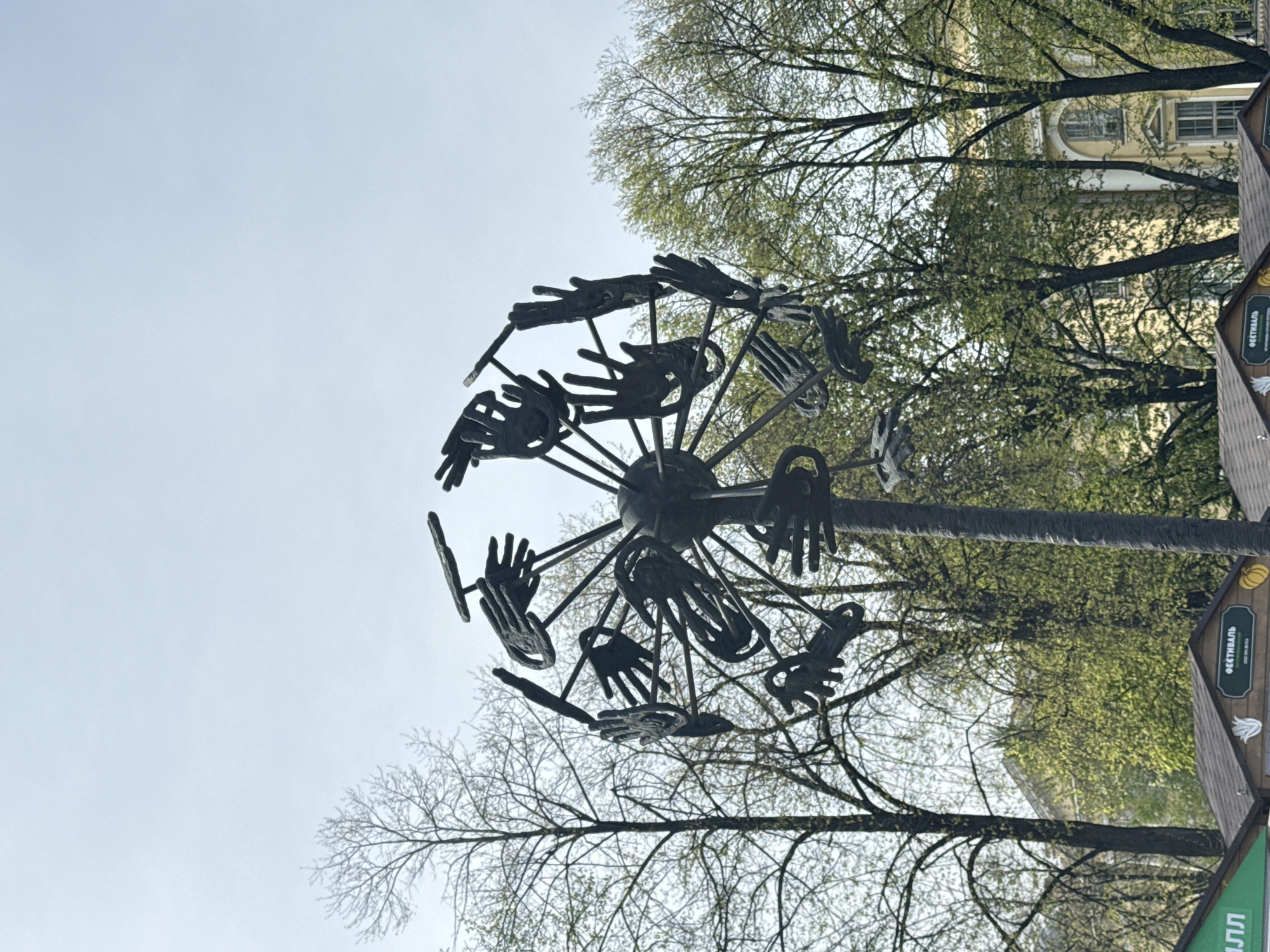 A metal sculpture featuring a wheel-like structure with hand-shaped extensions is positioned against a backdrop of trees and a partly cloudy sky. The sculpture appears to be in a public space, as evidenced by the presence of booth-like structures with inscriptions in Russian. A light-colored building is visible through the trees in the background. The overall atmosphere suggests a city park or urban setting in Saint Petersburg, Russia.