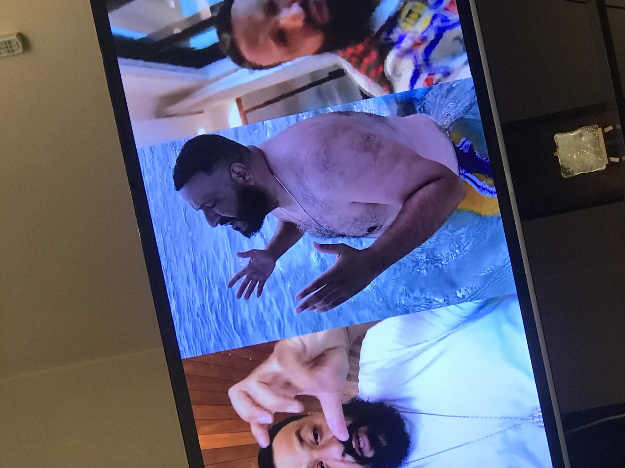 The image shows a TV screen displaying a compilation of clips featuring DJ Khaled. In the top clip, he is wearing a patterned shirt. The middle clip shows him shirtless in a pool of water. In the bottom clip, he is wearing a white shirt. The surrounding environment appears to be a room with neutral-colored walls and dark furniture. The location is not Yerevan, Armenia.