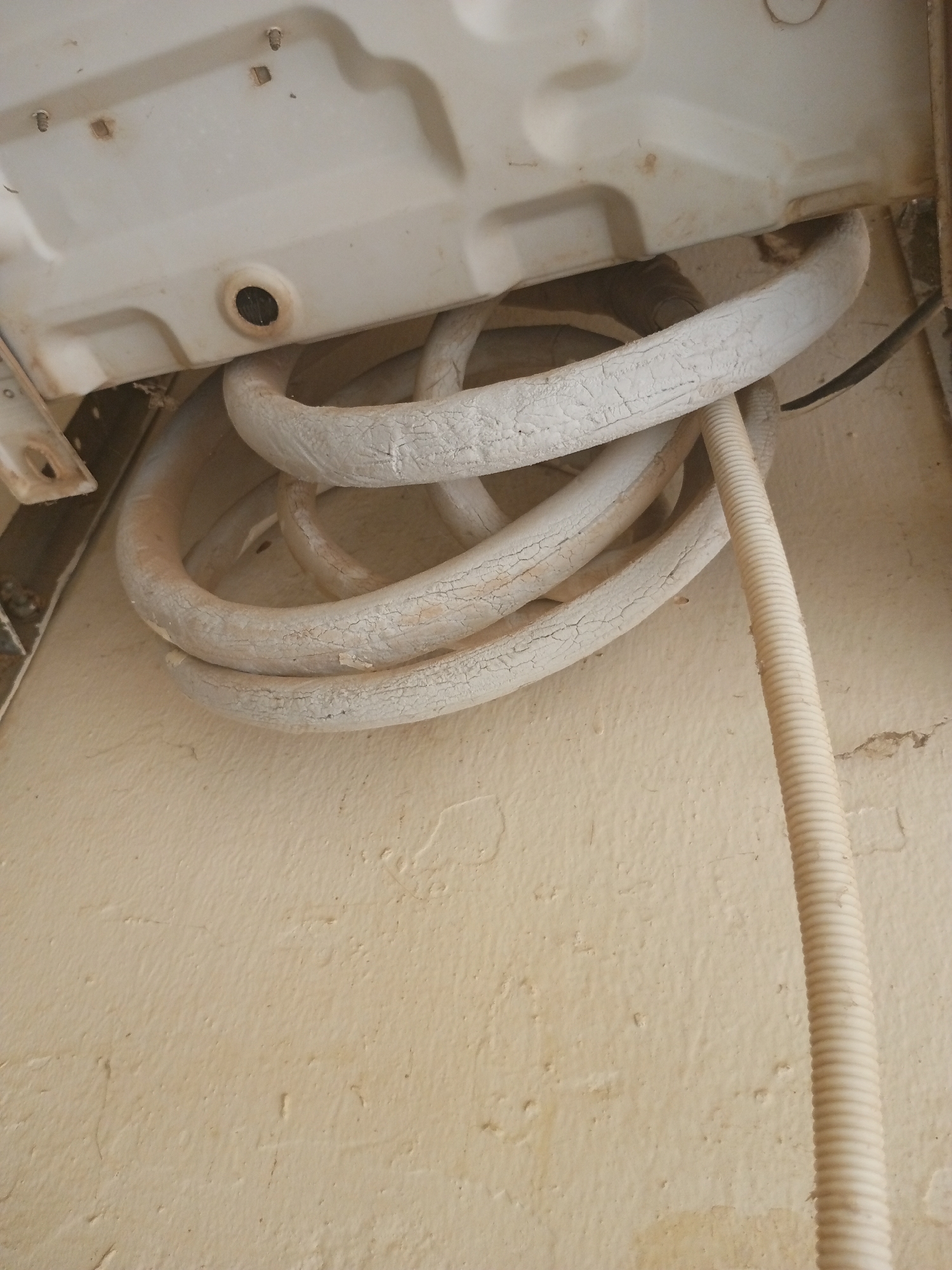 The image shows a close-up of an air conditioner unit's outdoor components, specifically the insulated copper tubing and a drainage hose. The tubing, coiled and white, appears weathered with visible cracks in its protective insulation. A ribbed, off-white drainage hose runs vertically down the right side of the frame. The background is a textured, cream-colored wall, likely stucco or concrete, common in outdoor settings. The lighting suggests it could be daytime, with shadows not being particularly harsh. There are no people visible, and no text is present in the image. The scene appears to be an outdoor installation of an air conditioning unit, possibly in a residential or commercial building in Jalingo, Nigeria. The overall impression is one of a functional, albeit somewhat aged, piece of external equipment.