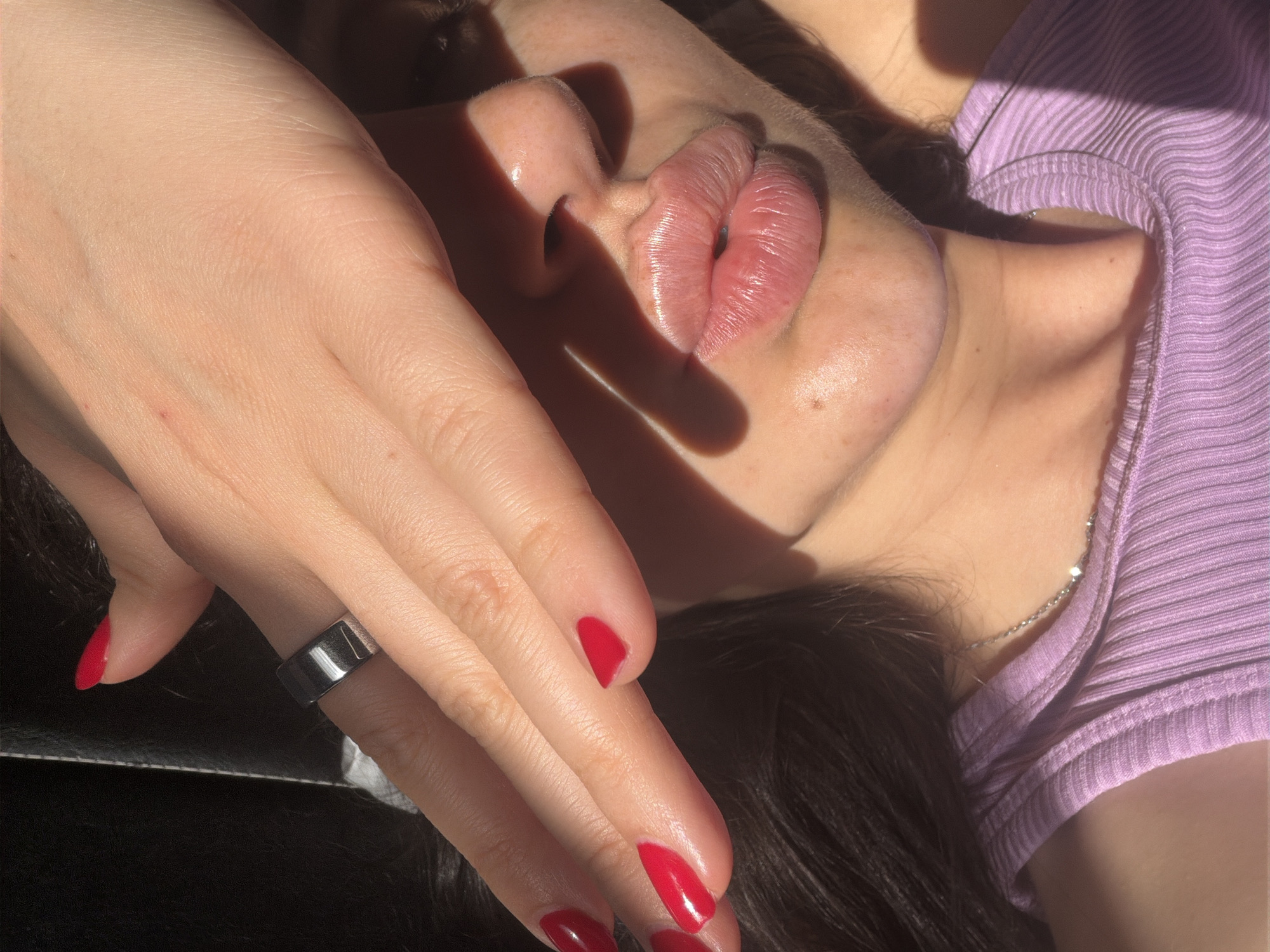 This close-up image captures a person's upper body, emphasizing their hand, lips, nose, and shoulder, illuminated by strong, direct sunlight. The scene is presented from a high-angle perspective, creating an intimate and somewhat abstract composition.
The primary subject's hand, positioned prominently in the foreground, features neatly manicured nails painted with a vibrant red polish. A sleek, dark-toned band ring, possibly silver or black, is worn on the ring finger. This hand casts a dramatic, sharp shadow across the person's face, partially obscuring the nose and mouth.
Visible facial features include full, glossy lips that appear slightly parted, and the bridge and tip of the nose, both highlighted by the intense sunlight. The skin texture is visible, showing natural details and a dewy appearance, particularly on the lips and around the nose. Dark hair frames the bottom and right side of the frame.
The person is wearing a ribbed, light purple top, visible on the right shoulder and chest area. A delicate silver-colored necklace chain is also discernible against the skin.
The strong sunlight creates a dramatic interplay of light and shadow, suggesting the photograph was taken during a sunny part of the day, likely outdoors or near a window. The sharp shadows and bright highlights contribute to the overall aesthetic, emphasizing contours and textures. The setting is inferred to be in Budva, Montenegro. There is no visible text in the image.