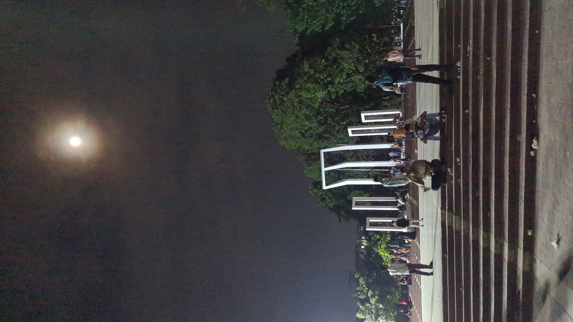 The image depicts a night scene at the Aparajeyo Bangla monument in Dhaka, Bangladesh. The sky is dark with a bright full moon visible, casting an ethereal glow. In the foreground, a wide set of concrete stairs leads up to the monument, which is comprised of several tall, white, abstract structures. Several individuals are present on the stairs and around the monument.
In the mid-ground, three young men are sitting on the stairs. One is seated cross-legged, looking towards the left, with a light-colored shirt and darker pants. Another is also seated with legs bent, wearing a light-colored shirt and blue jeans, and looking down. A third man stands to the right of these two, wearing a dark blue shirt and dark pants, looking towards the left. Further back, near the monument, a man in a white shirt and dark pants stands facing left. Other figures are scattered throughout the background, some appearing to be sitting or standing.
The environment is illuminated by artificial lighting, which highlights the white structures of the monument and casts shadows. Trees with dense foliage are visible behind the monument, suggesting a park-like setting. The stairs appear somewhat worn, and there are some pieces of litter visible on them, indicating public use. The overall atmosphere is calm and social, with people gathering in a public space at night.