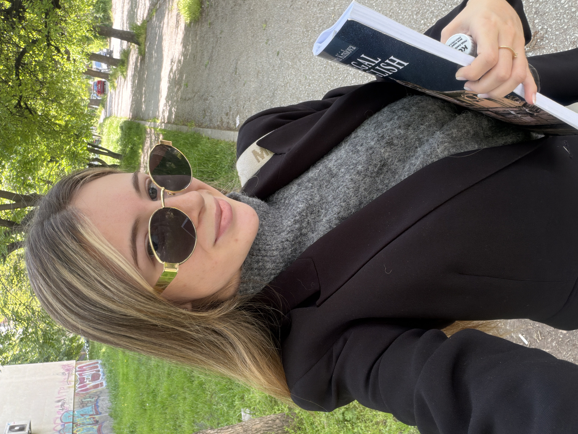 A woman with blonde hair wearing gold-framed sunglasses is standing outdoors holding a book titled "LEGAL ENGLISH" by Maria Kristova. She is wearing a black blazer over a gray turtleneck sweater. The background shows a city park scene with trees and a paved path. Graffiti is visible on a wall in the foreground. The location is within the city of Plovdiv, Bulgaria.