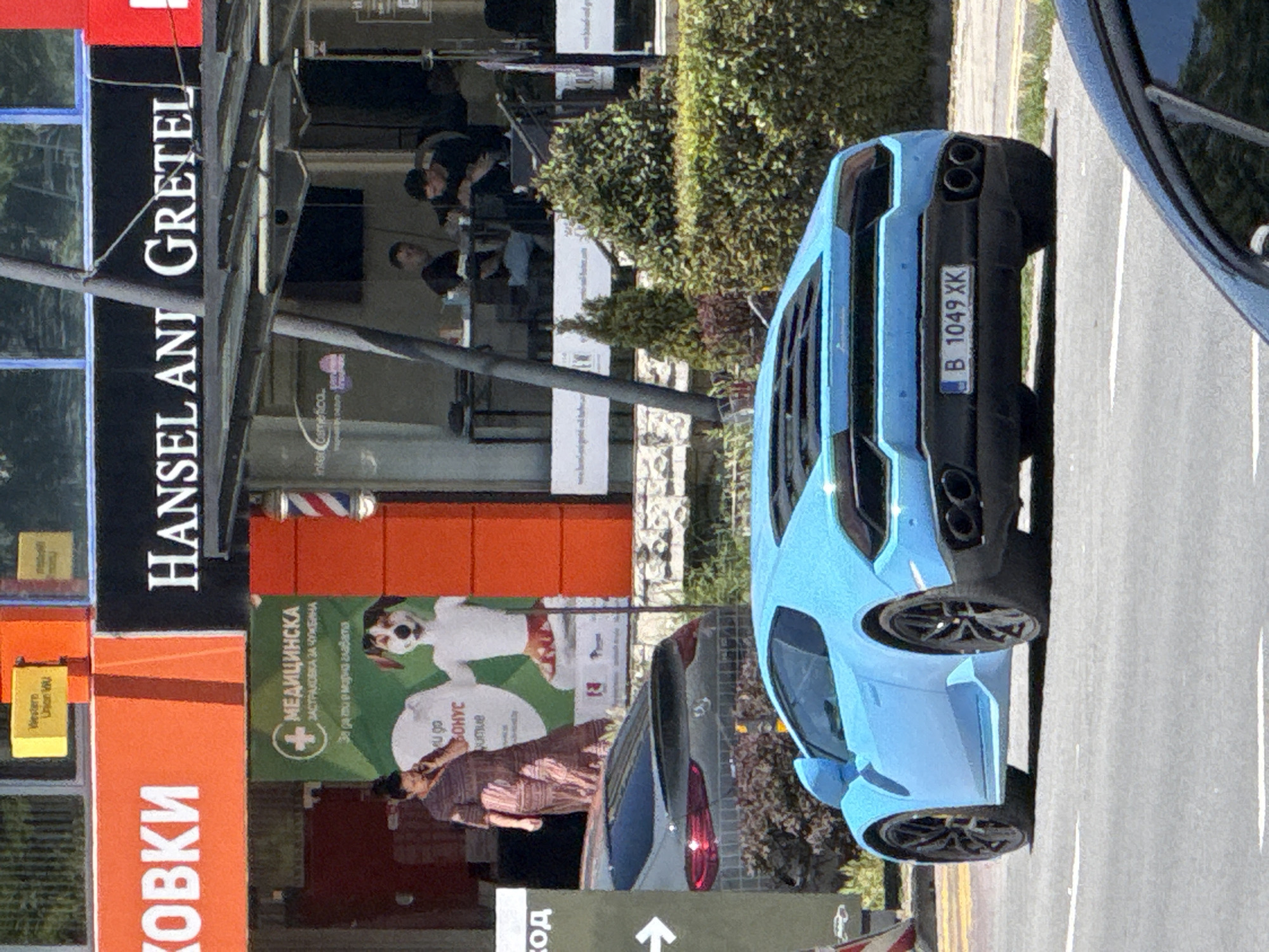 The image shows a street scene in the city of Varna, Bulgaria. In the foreground, a bright blue Lamborghini sports car is parked on the right side of the road. The car has black wheels and a license plate that reads "B 1049 XK". Behind the Lamborghini, a dark-colored car is partially visible.
The background features a row of shops and businesses. One shop has a large orange sign with white lettering that appears to say "КОВКИ". Next to it is a black sign with white lettering that reads "HANSEL AND GRETEL". A woman in a patterned dress is standing in front of a green banner with text and an image of a dog. Further back, several people are seated at tables outside a cafe or restaurant under a metal awning. The environment appears to be urban, with buildings, signage, and street infrastructure visible.
