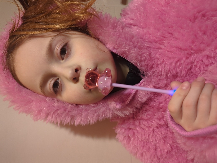 A young child, likely female, with light brown hair and fair skin, is depicted in a close-up portrait. She wears a pink, hooded, fuzzy coat, with the hood framing her face. The child holds a translucent pink, teddy bear-shaped object, resembling a lollipop or toy, on a light pink stick. The object is positioned in front of her mouth. Her right hand, partially visible, grips the stick, which emits a blue light from its base. The child maintains direct eye contact with the camera. The background consists of a plain, light-colored wall, offering no discernible identifying features for a specific location within Moscow, Russia.