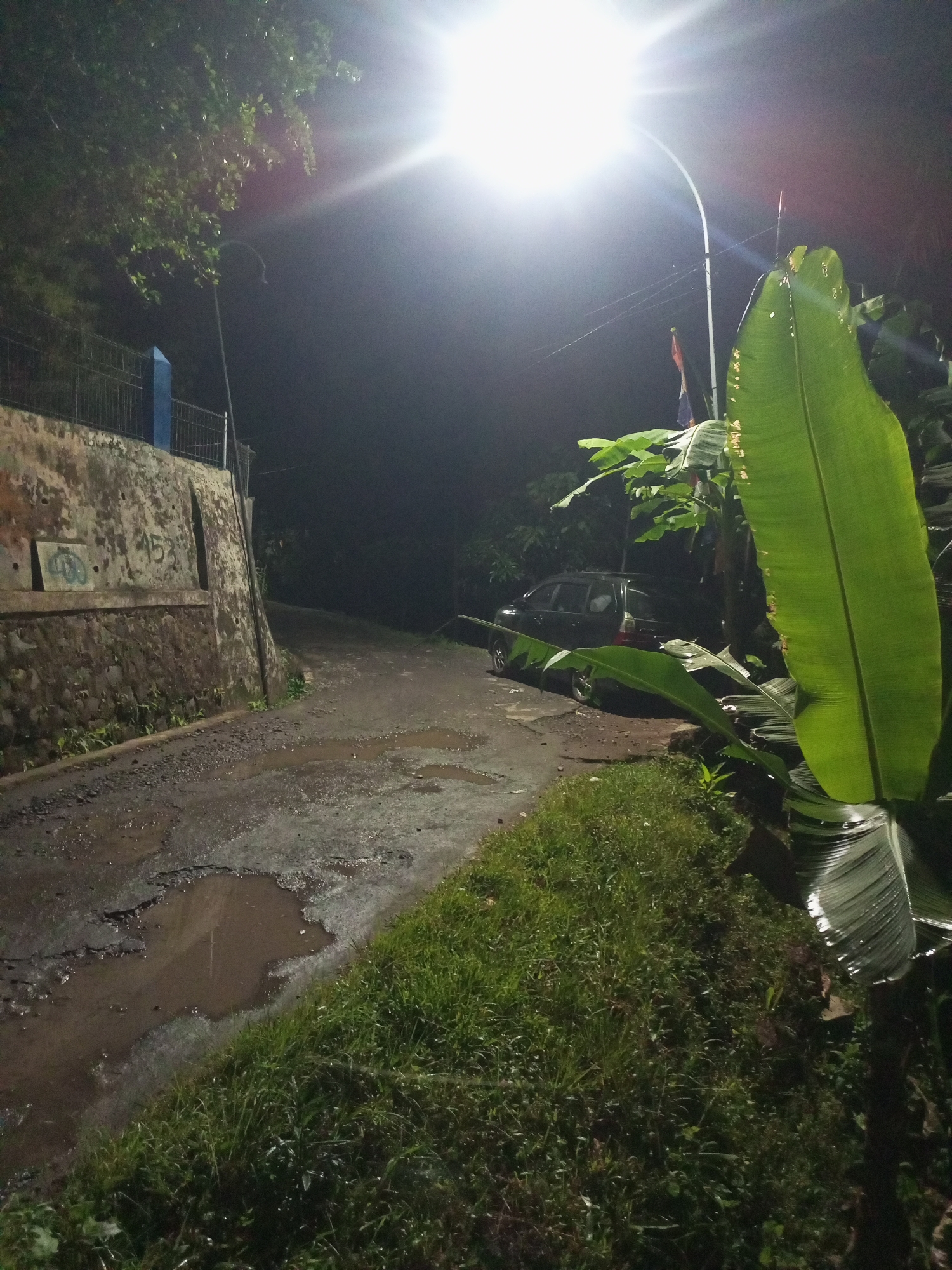 This is a nighttime photograph taken on a rural road in Cidahu, Indonesia. A bright street lamp illuminates the scene, casting a strong glare. To the left, a stone wall with some graffiti runs alongside the road. The road itself is wet and appears to be in disrepair, with several potholes filled with muddy water. Lush green grass and large banana leaves dominate the right side of the frame. In the background, a dark-colored car is parked on the side of the road, partially obscured by vegetation. The overall impression is of a dimly lit, somewhat neglected area.