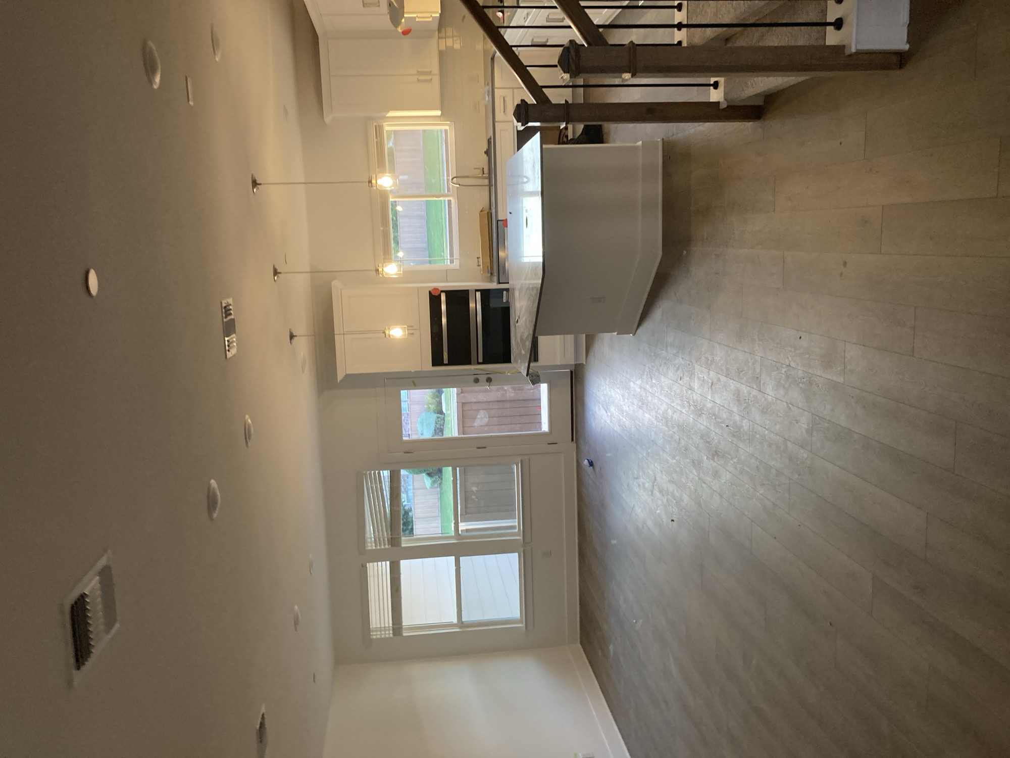 The image depicts an interior scene of a modern, unfinished residential space, likely a new construction, in Round Rock, United States of America. The room appears to be an open-concept living and kitchen area.
In the foreground, a light-colored, wood-plank style flooring covers the ground. To the right, a wooden staircase with black balusters ascends, suggesting a multi-story home.
The middle ground features a white kitchen island with a granite countertop. Behind the island, a kitchen area is visible with white cabinets, a stainless steel sink, and a faucet. Two ovens are stacked vertically, one above the other, to the left of the kitchen counter. Pendant lights hang over the island. A large window with horizontal blinds overlooks a backyard with a fence and greenery. A glass-paneled door, likely an exterior exit, is also visible to the left of the window.
The background shows a white wall on the left and the continuation of the kitchen cabinetry and appliances. The ceiling is white and features recessed lighting and a rectangular air vent.
The space appears to be empty of people and is in a state of construction or renovation, with some debris visible on the floor. The lighting is artificial, casting shadows and highlighting the features of the room.