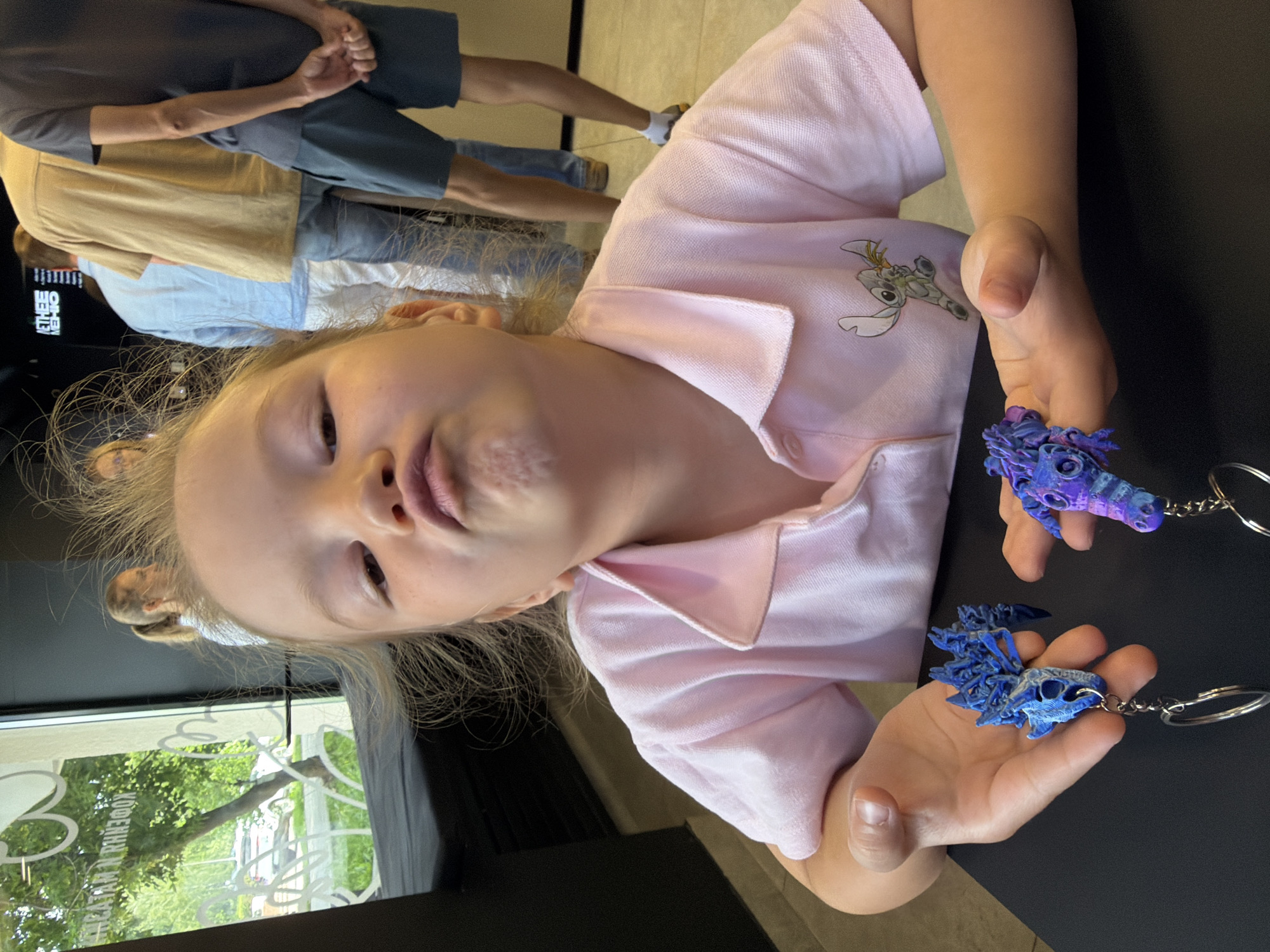 A young Caucasian girl, appearing to be around 4-6 years old, with blonde hair and light skin, makes a puckered face at the camera. She is wearing a pink polo shirt with a Stitch graphic on the chest. Her hands are open, displaying two small, intricately designed 3D-printed dragon skull keychains. One keychain is predominantly blue, and the other is purple and blue. The girl is seated at a black table, with her hands and the keychains in the foreground. In the background, out of focus, several adults are standing in what appears to be a queue. Through a window to the left, trees and a white car are visible, suggesting an outdoor scene behind the establishment. The overall setting seems to be an indoor public space, possibly a cafe or shop, given the presence of people queuing and a visible sign in the background with Cyrillic text that is partially obscured. A sign in the background that is mostly visible has the text "ЕТНЕЕ МЕНЮ" which translates to "ETHEE MENU". There is also some text on the window in Cyrillic that is partially obscured and appears to be advertising "КОФЕ И НАПИТКИ" which translates to "COFFEE AND DRINKS". The image was taken in Izhevsk, Russia.