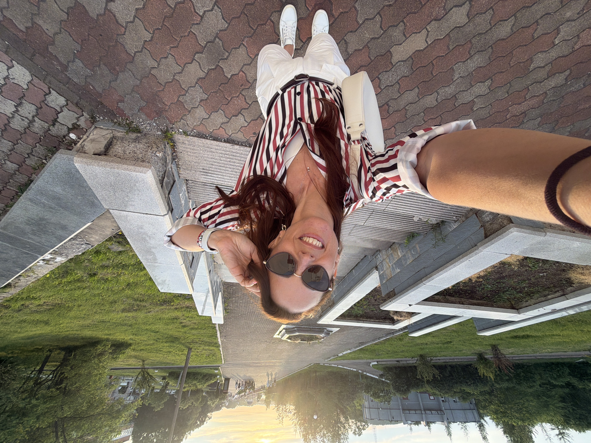 A woman is taking a selfie in a park in Birobidzhan, Russia. She is wearing a striped shirt, white shorts, and white sneakers. She has sunglasses on and is holding her hair. The park has paved walkways, grass, and trees. There is a building in the background. The sun is setting.